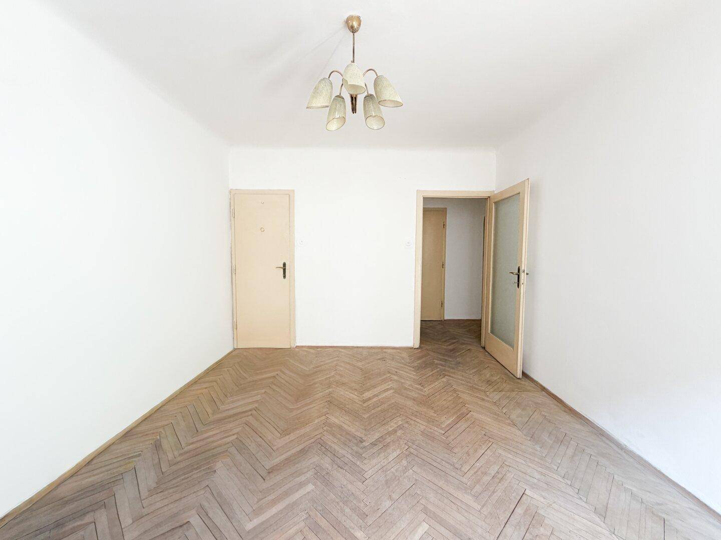 Leeres Zimmer mit Fischgrätparkett und zwei Türen, die zu verschiedenen Bereichen der Wohnung führen.