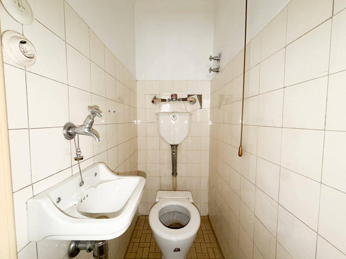 Kleiner Toilettenraum mit Waschbecken und WC, umgeben von weißen Fliesen und einem hellbraunen Boden.