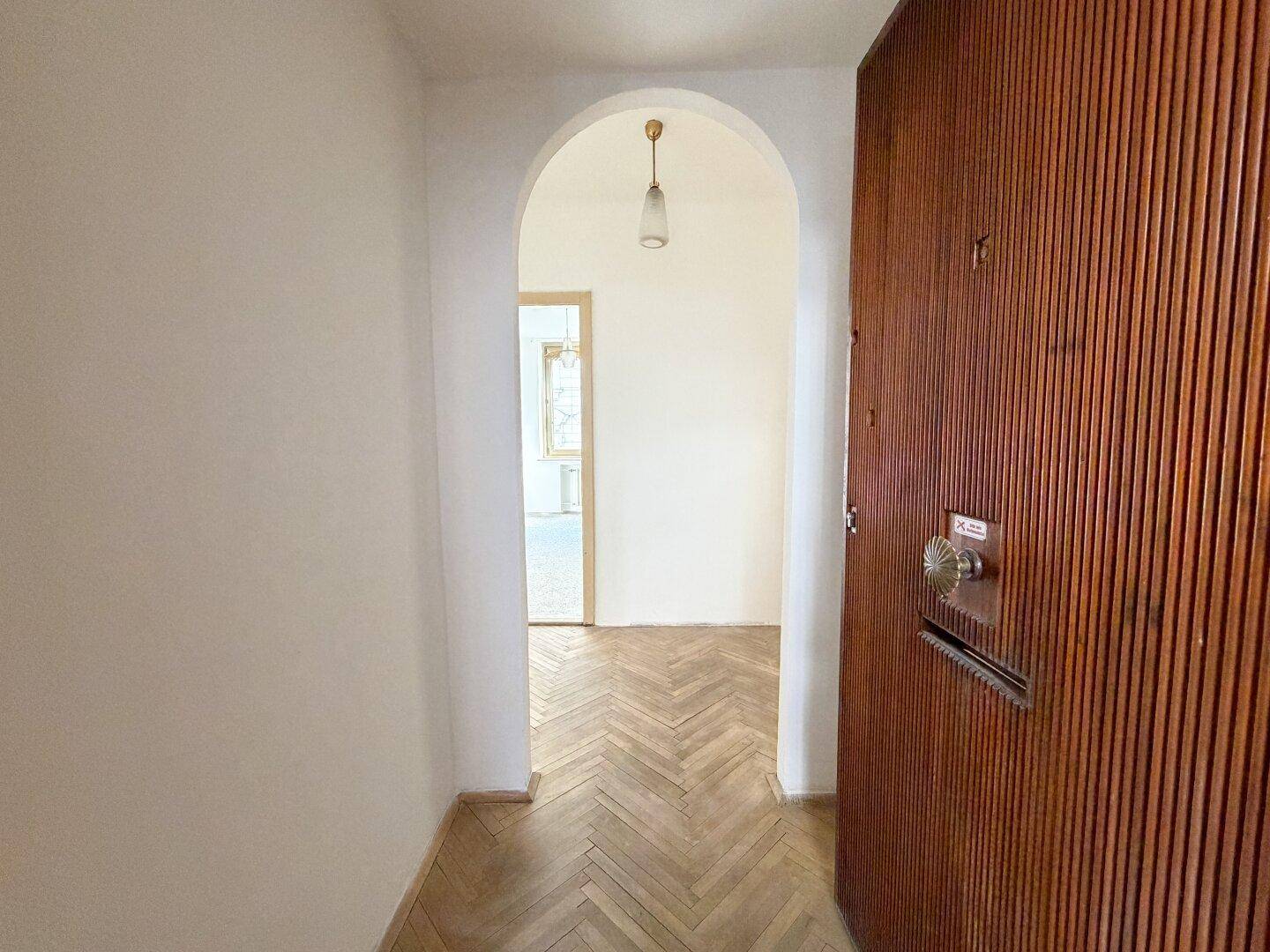 Eingangsbereich mit Holztür, Fischgrätparkett und einem eleganten Rundbogen, der in den nächsten Raum führt.