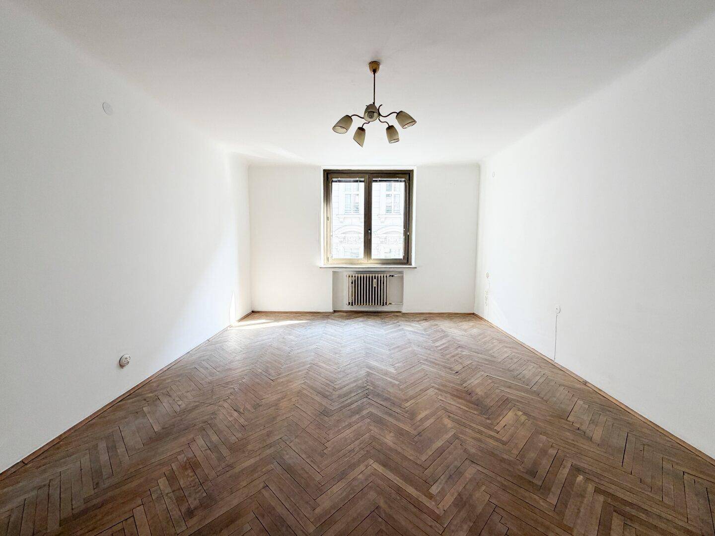 Geräumiges leeres Zimmer mit Fischgrätparkett, weißen Wänden und einem Fenster mit Heizkörper.