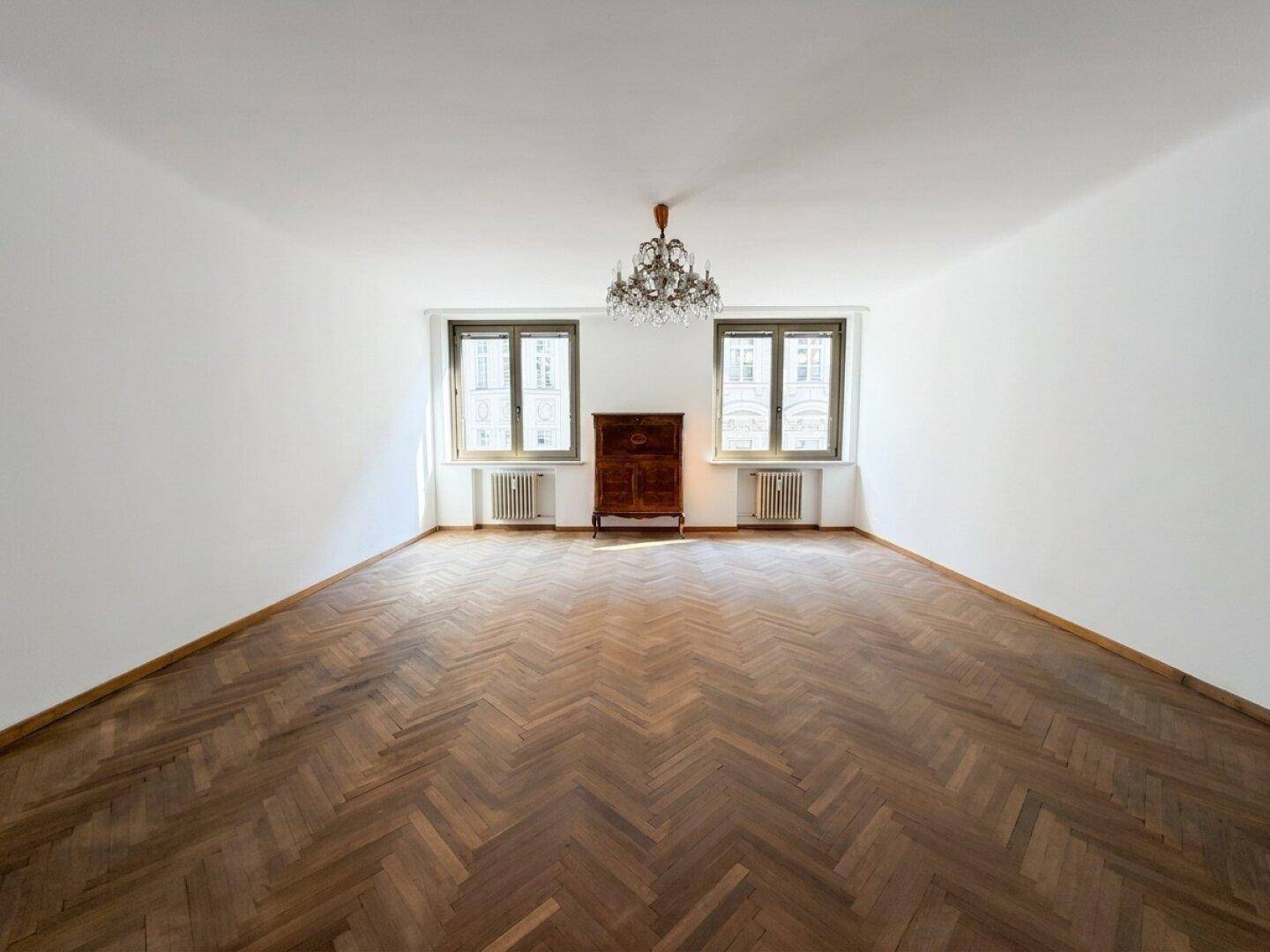 Geräumiges Zimmer mit Fischgrätparkett, zwei Fenstern und einem eleganten Kronleuchter.