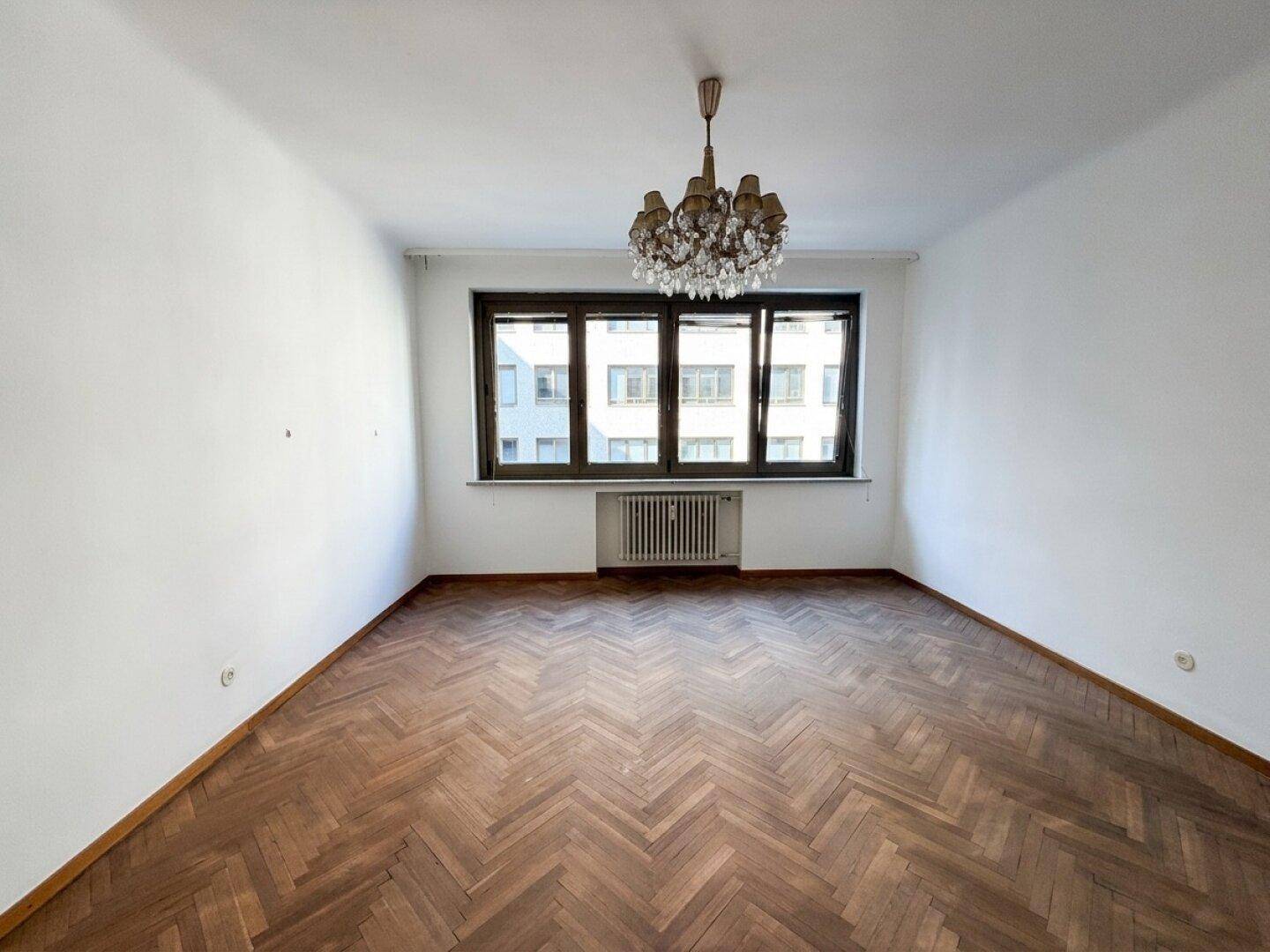 Leeres Zimmer mit Fischgrätparkett, großem Fenster und einem dekorativen Kronleuchter.