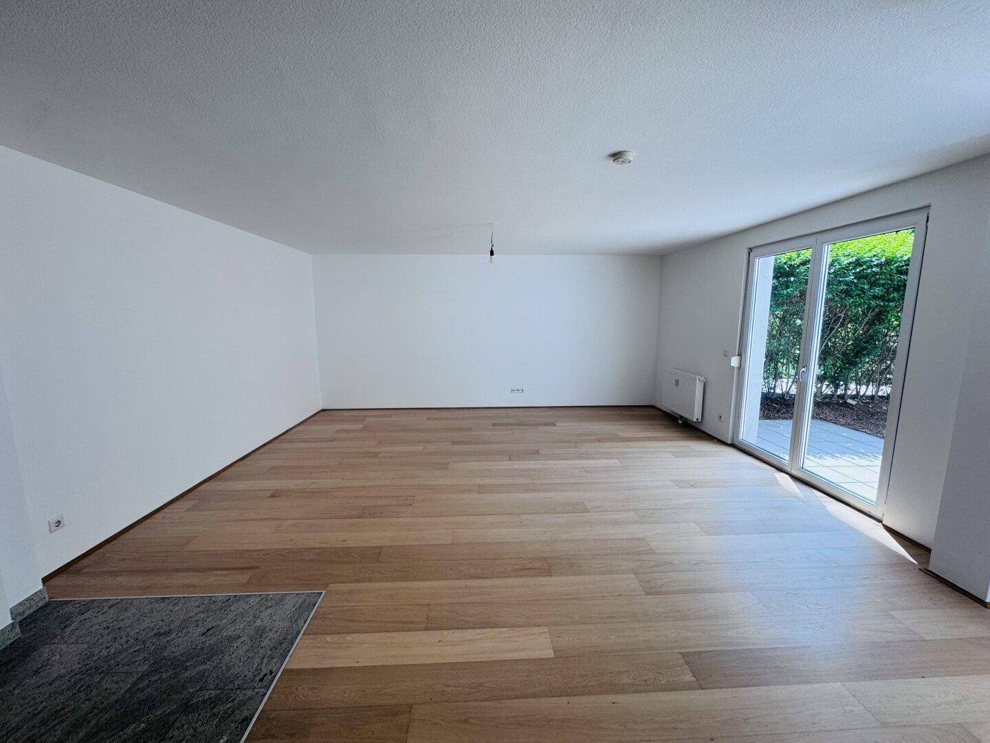 Helles, leeres Wohnzimmer mit hochwertigem Holzfußboden und großer Glasschiebetür zum Außenbereich.