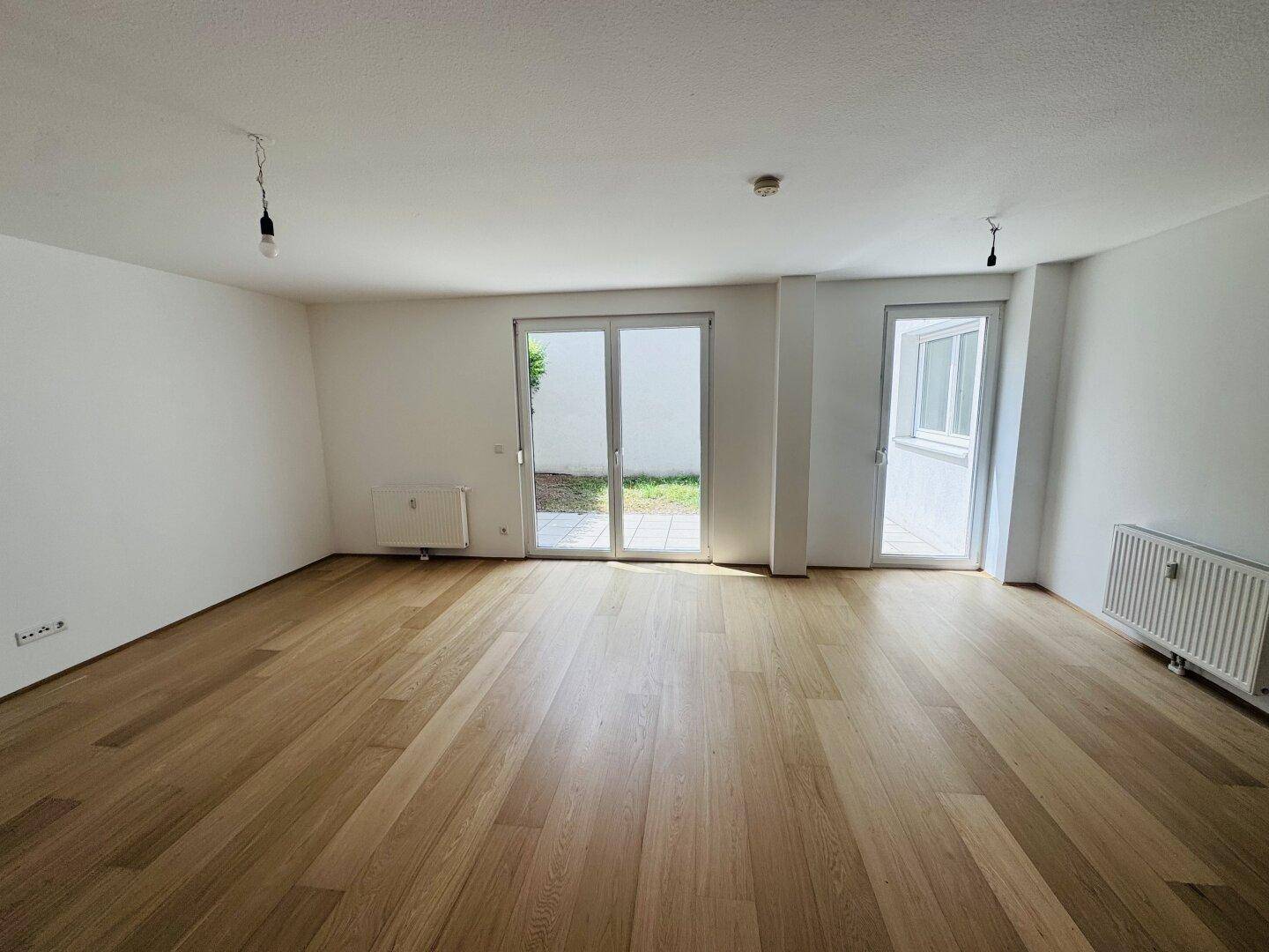 Geräumiges Wohnzimmer mit hellem Holzfußboden und zwei Glastüren, die Zugang zum Garten bieten.