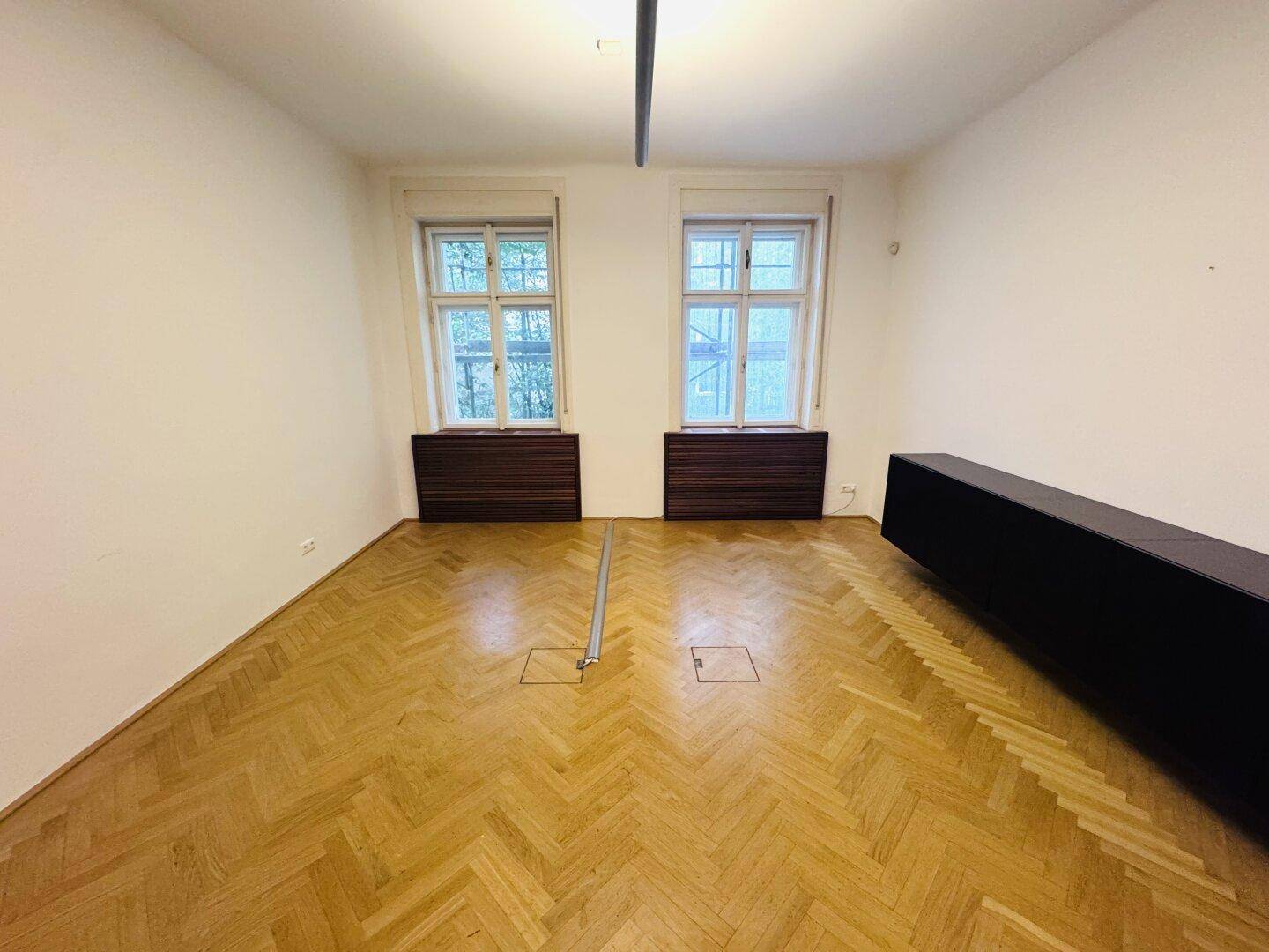 Heller Büroraum mit Fischgrätparkett, zwei Fenstern und stilvollen Heizkörperverkleidungen aus Holz.