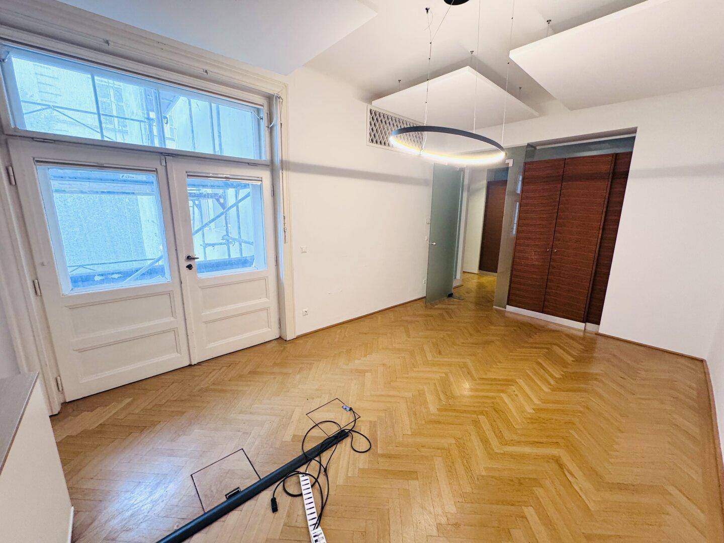 Heller Büroraum mit Fischgrätparkett, großen Doppeltüren und einer modernen Deckenleuchte.