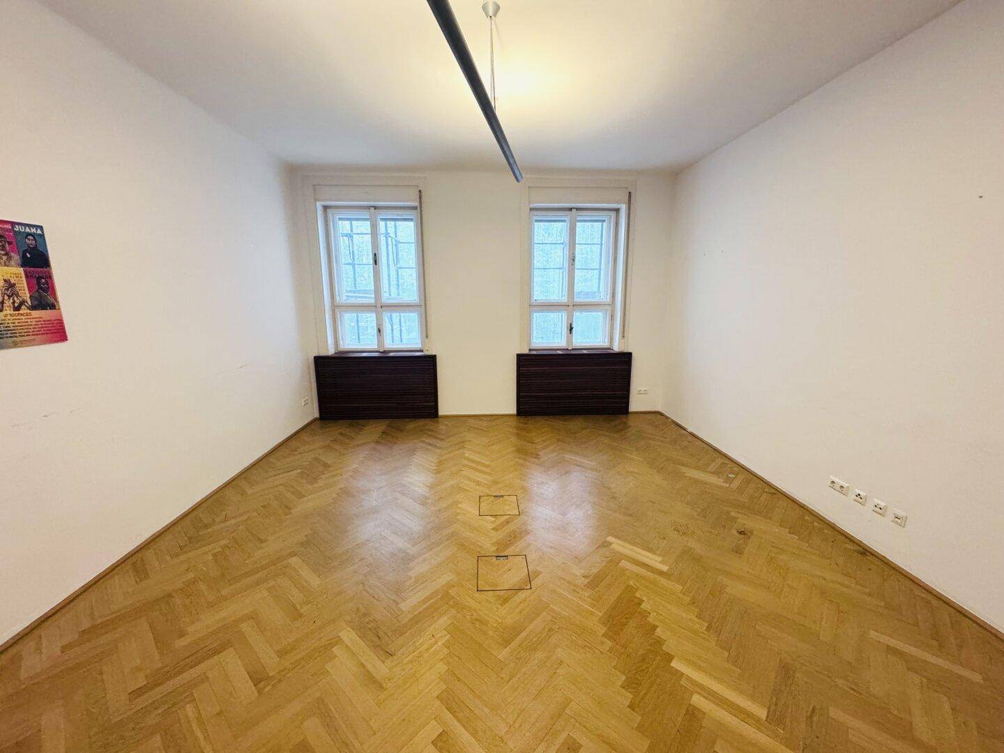 Heller Büroraum mit Fischgrätparkett, zwei Fenstern und dunklen Heizkörperverkleidungen.