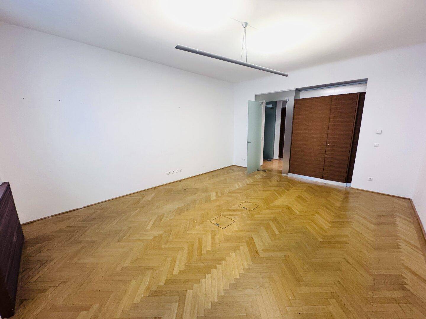 Büroraum mit Fischgrätparkett, weißen Wänden und integrierten Holzschränken im Hintergrund.