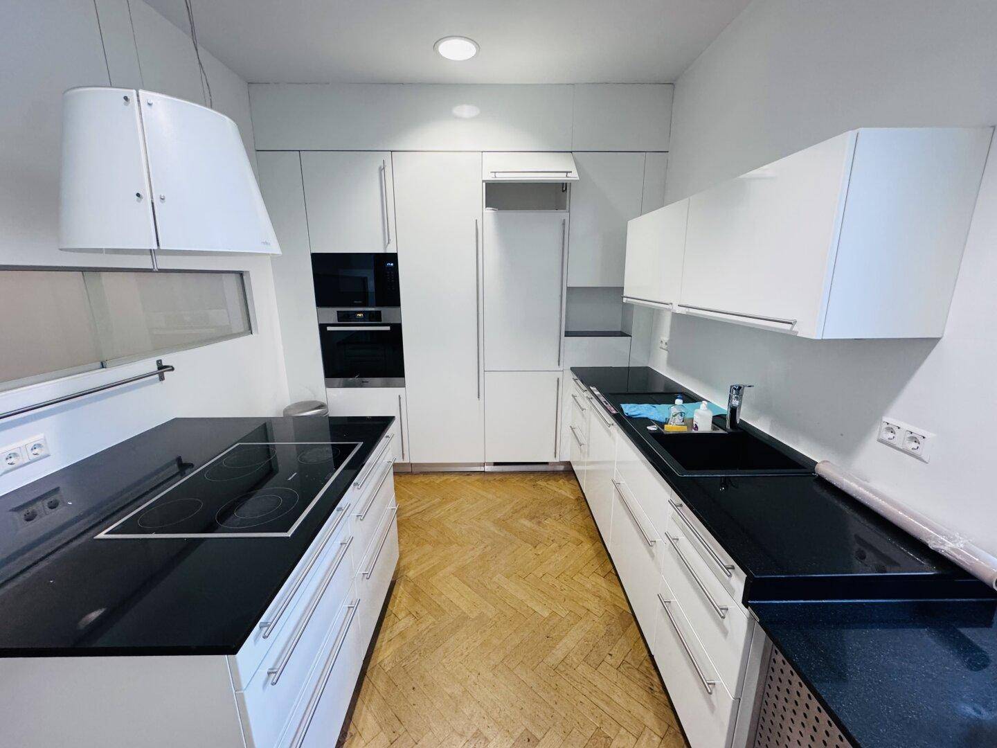Moderne Küche mit weißen Schränken, schwarzen Arbeitsplatten, Induktionskochfeld und Einbaugeräten.