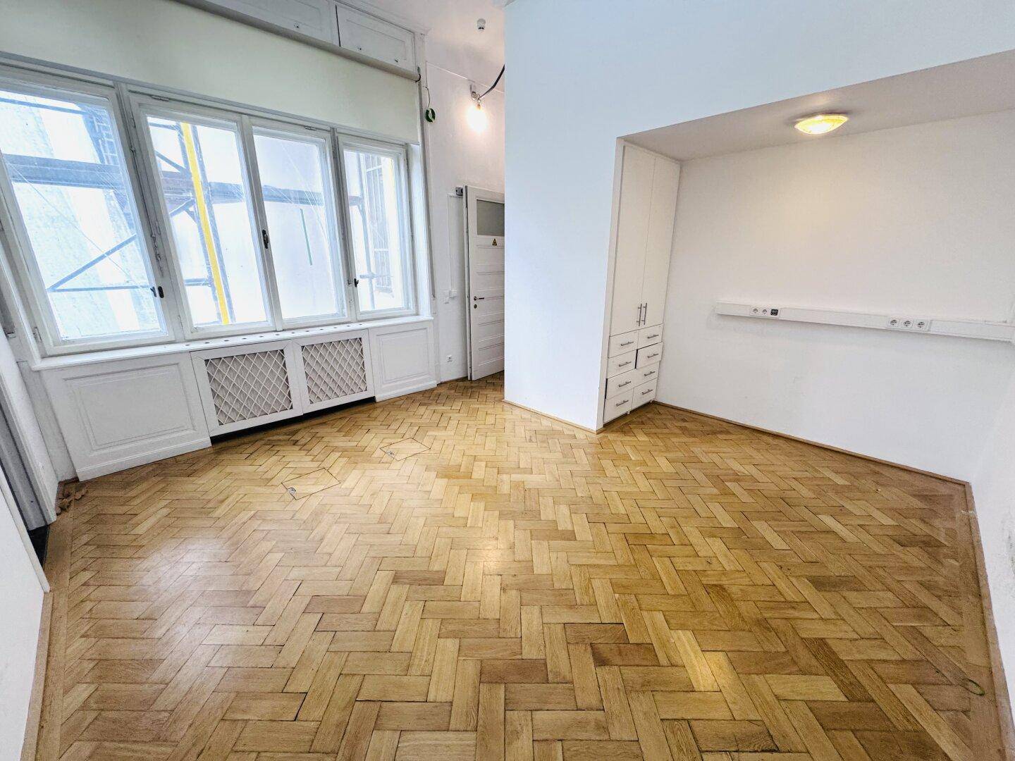 Heller Büroraum mit Fischgrätparkett, großen Fenstern und einem integrierten weißen Schrank.