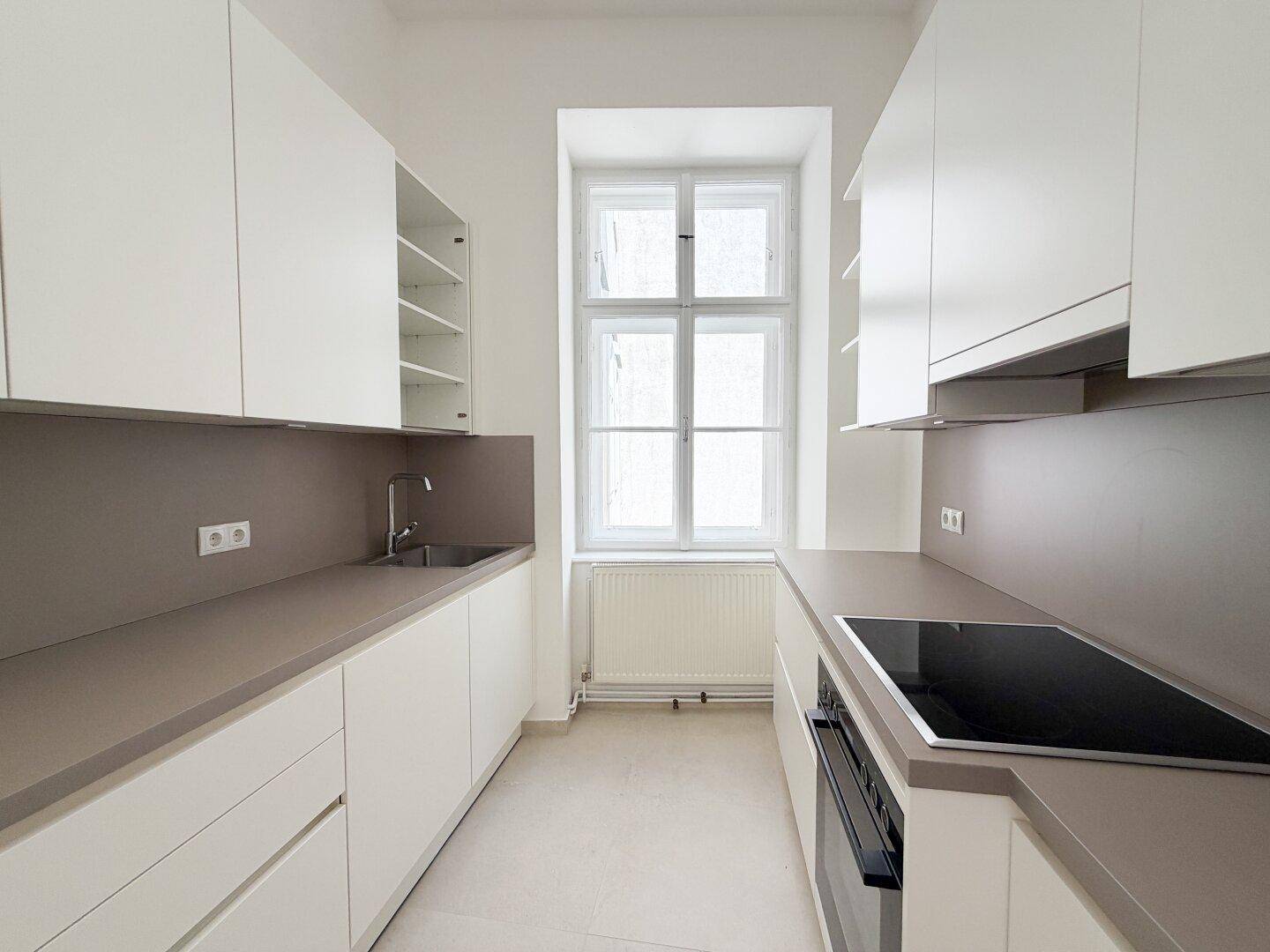 Moderne Einbauküche mit weißen Schränken, grauen Arbeitsplatten und Fenster für Tageslicht.