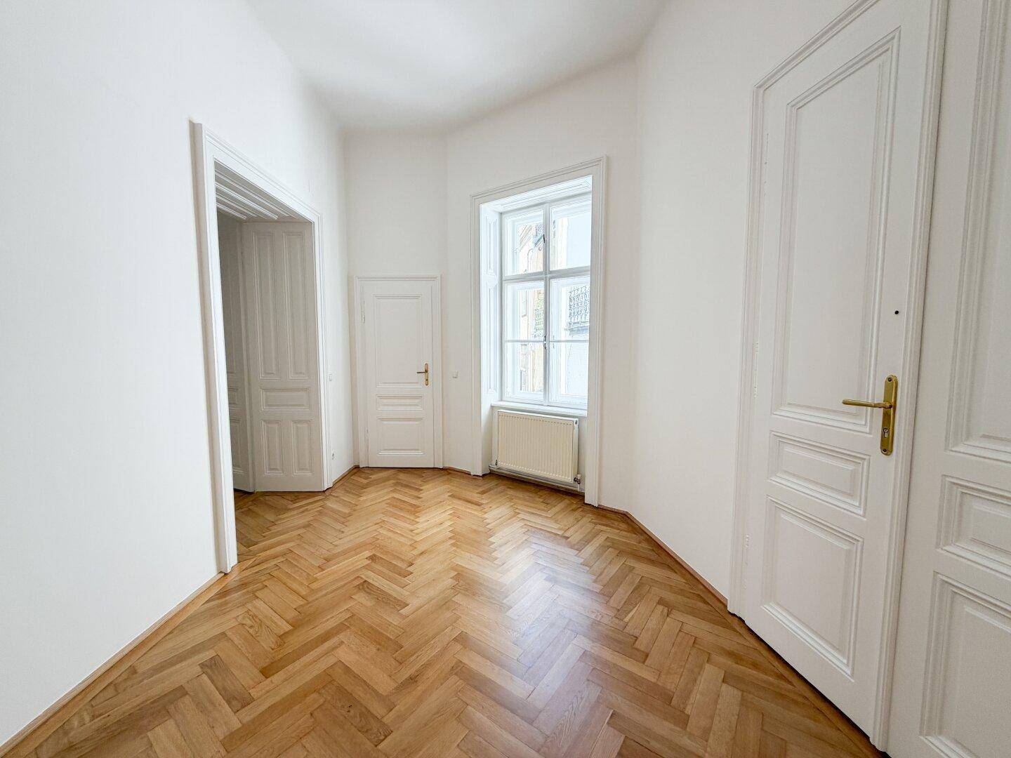 Eingangsbereich mit Fischgrätparkett, hohen Türen und einem Fenster, das für Helligkeit sorgt.