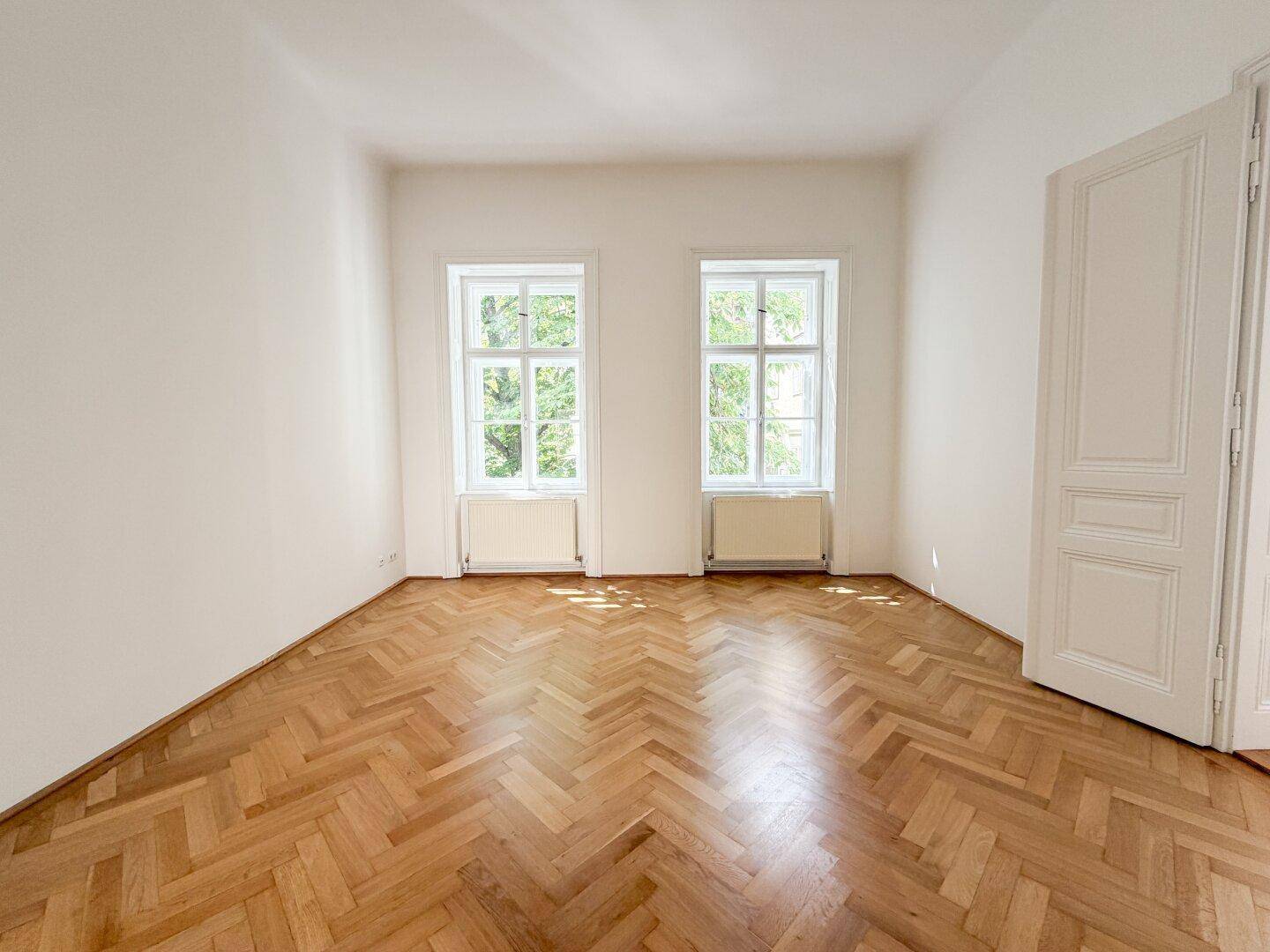 Geräumiges Zimmer mit klassischem Fischgrätparkett und zwei großen Fenstern für viel Helligkeit.
