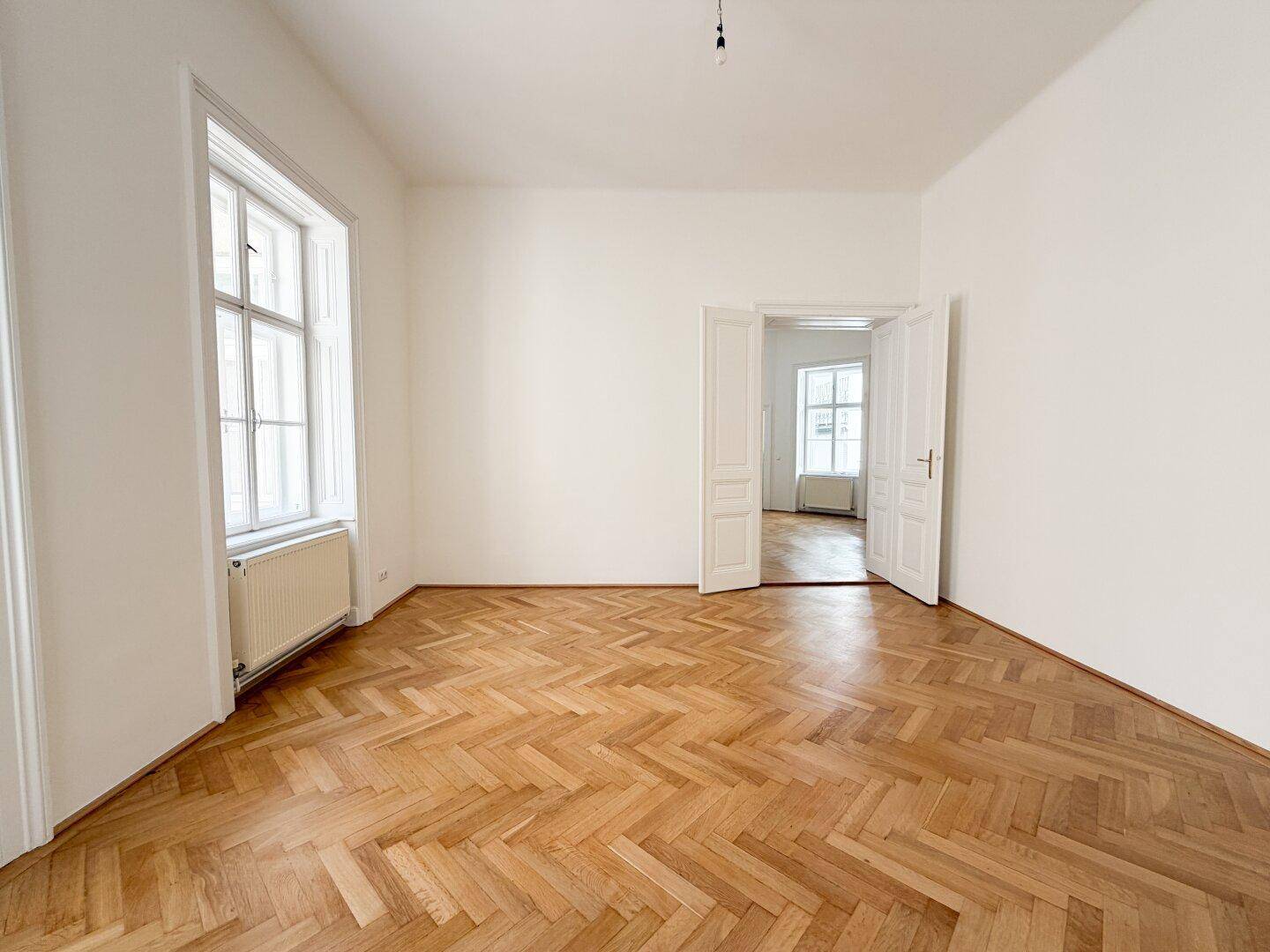 Helles Zimmer mit Fischgrätparkett und hohen Fenstern, die einen Blick ins Grüne bieten.