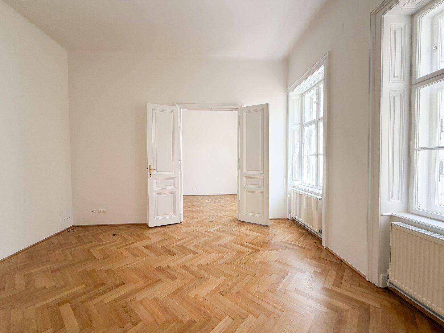 Geräumiges Zimmer mit Fischgrätparkett und hohen Flügeltüren, die zu einem angrenzenden Raum führen.
