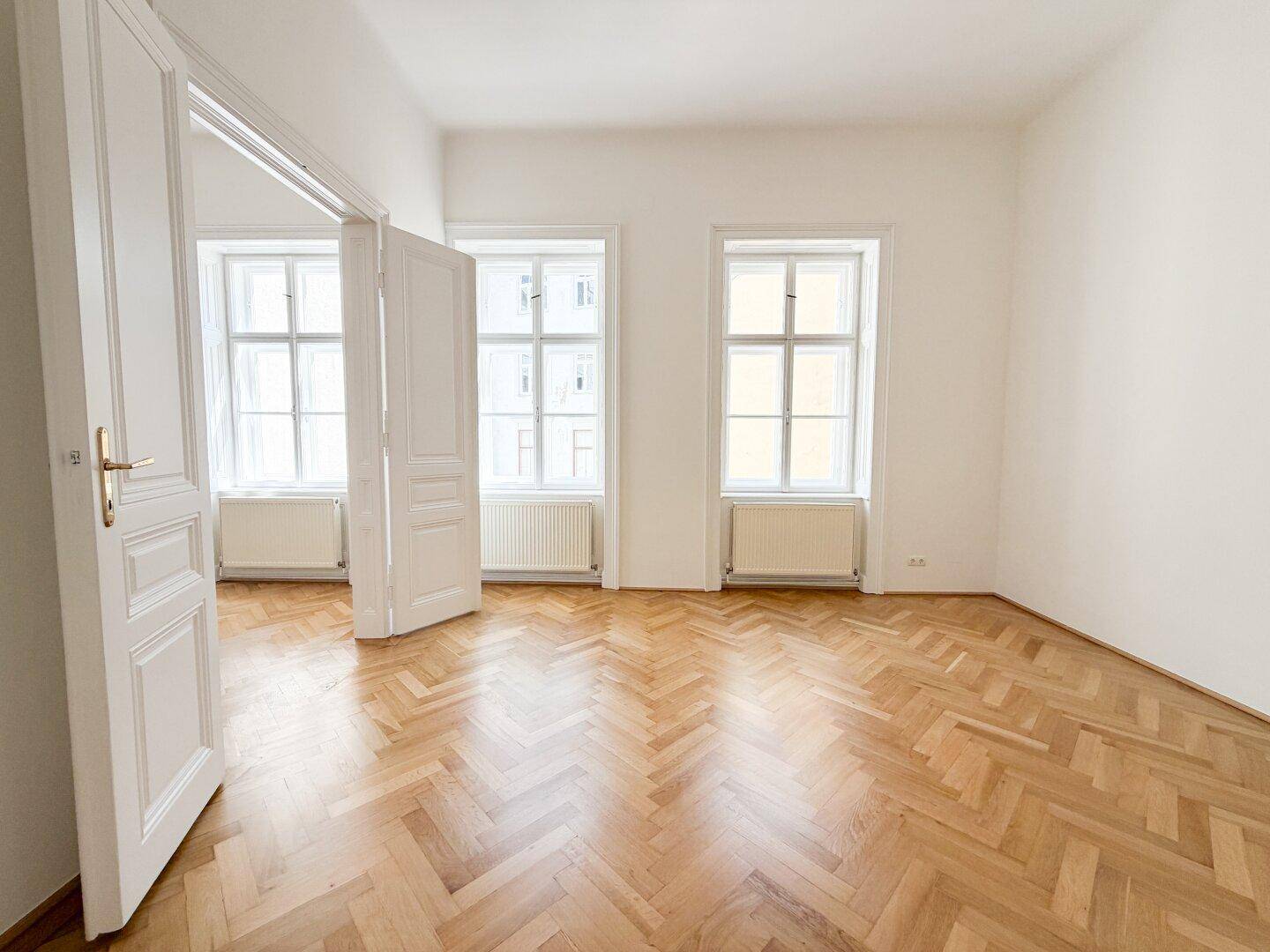 Großzügiger Raum mit Fischgrätparkett, hohen Fenstern und Flügeltüren, die zu einem weiteren Zimmer führen.