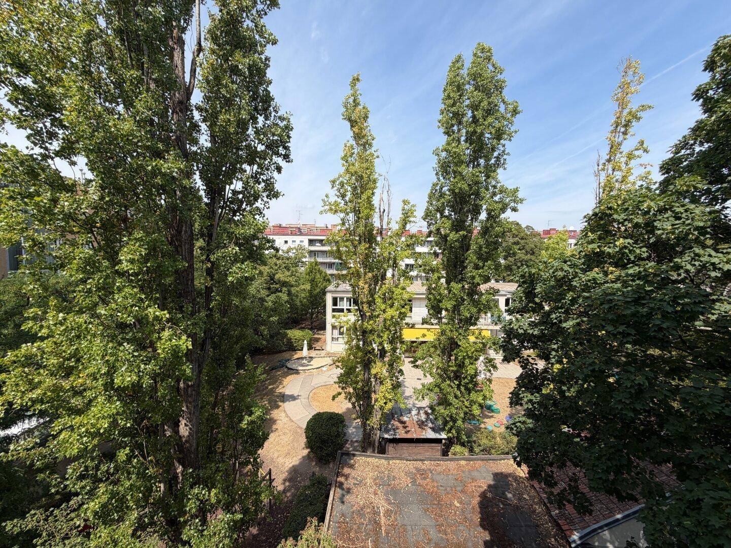 Blick von oben auf einen grünen Innenhof mit hohen Bäumen und umliegenden Gebäuden.