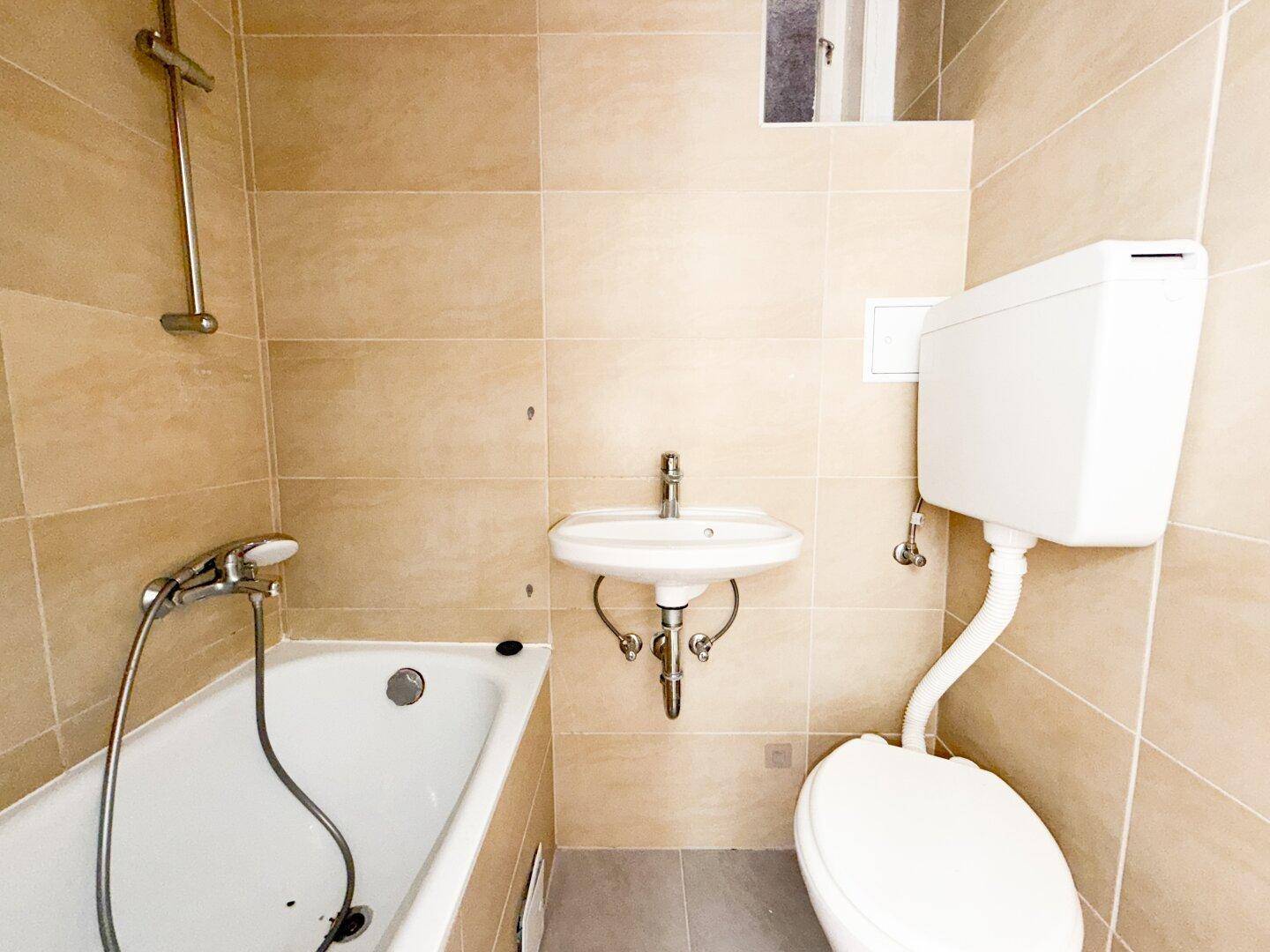 Funktionales Badezimmer mit beigefarbenen Fliesen, Badewanne mit Duschfunktion, Waschbecken und Toilette.