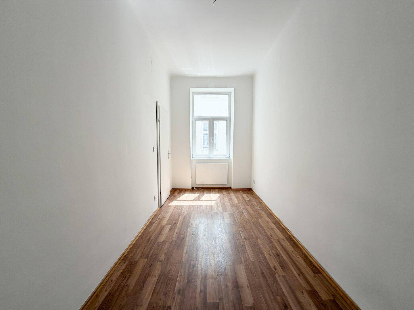 Schmales, helles Zimmer mit Laminatboden und einem Fenster, ideal als Schlaf- oder Arbeitszimmer.