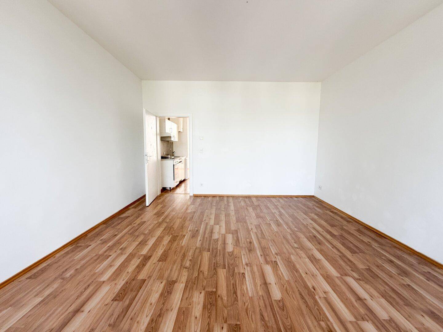 Geräumiges, leeres Zimmer mit Laminatboden und Zugang zu einer kleinen Küche.