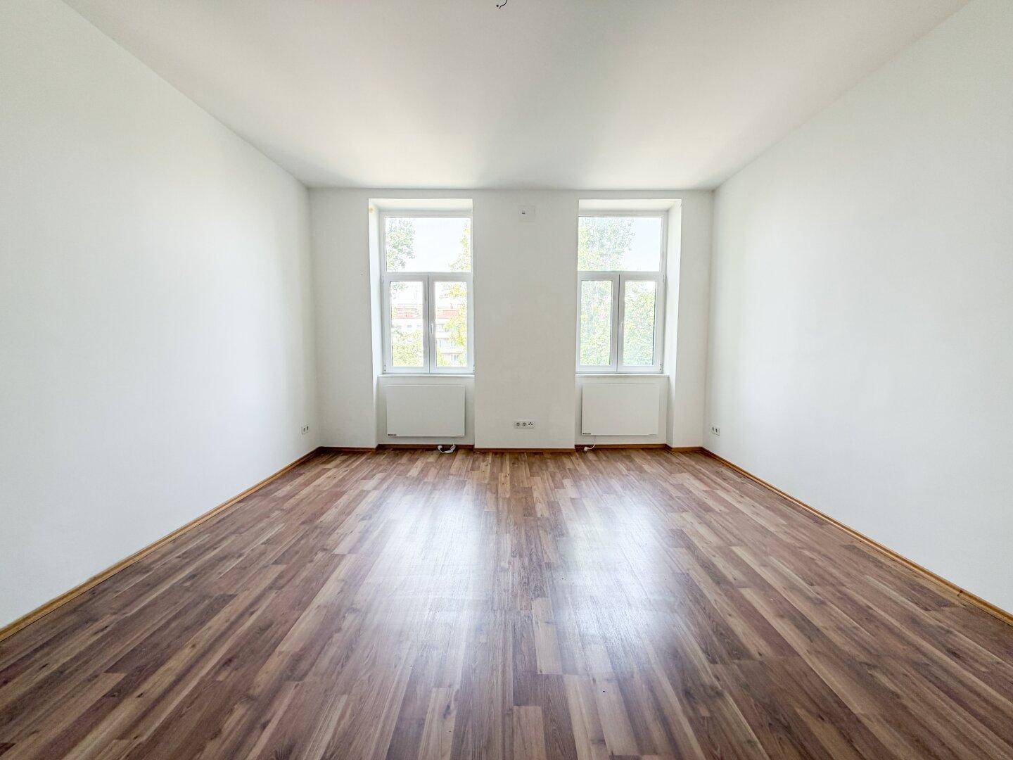 Helles, leeres Zimmer mit weißen Wänden und Laminatboden, ausgestattet mit zwei Fenstern.