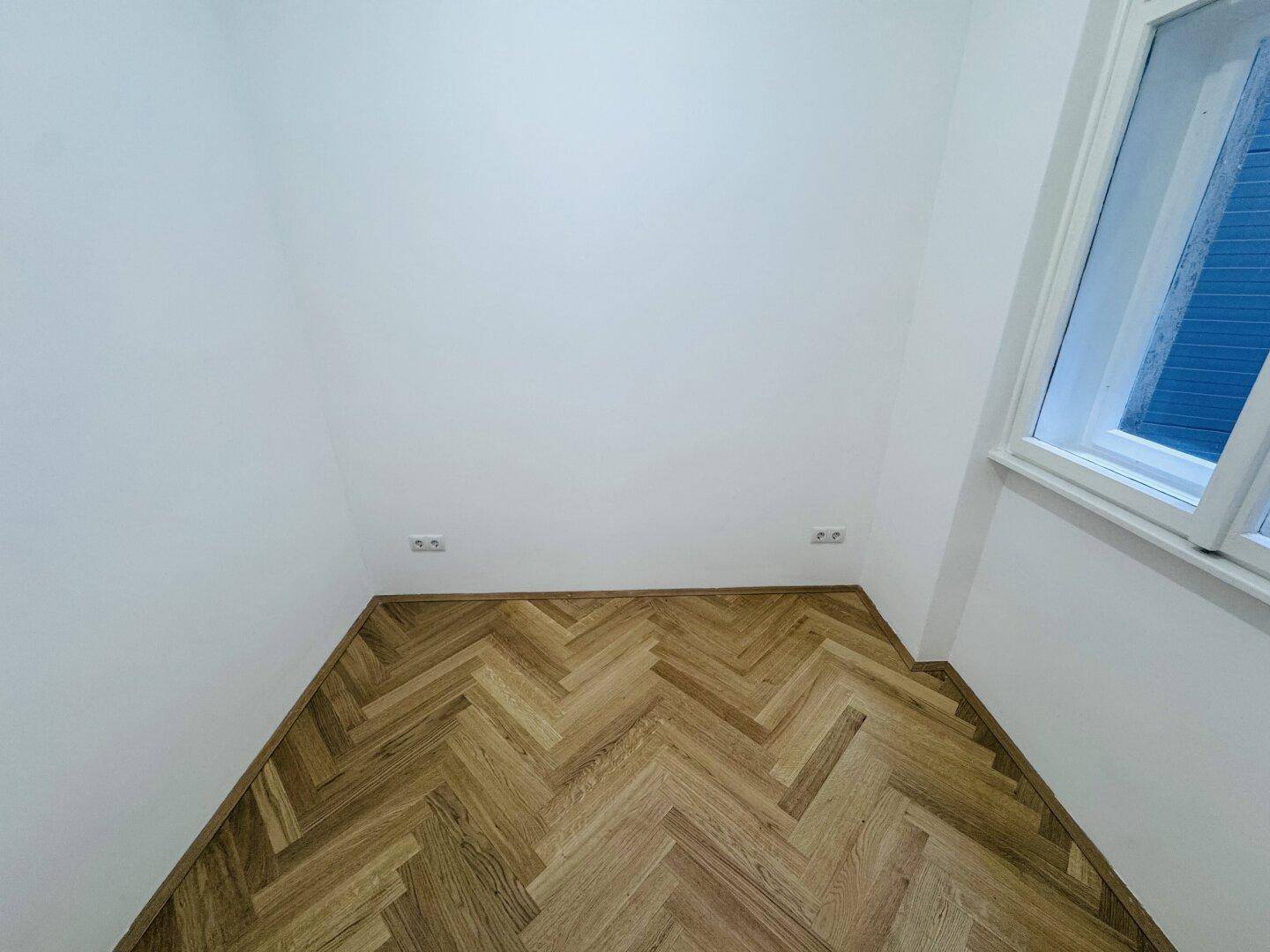 Kleiner, heller Raum mit Fischgrätparkett und einem Fenster, ideal als Abstellraum oder begehbarer Kleiderschrank.