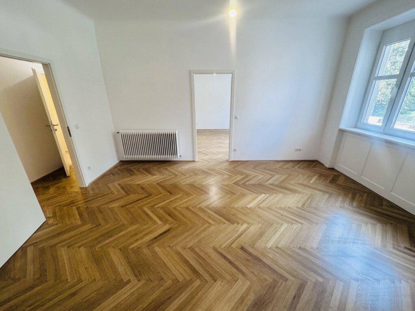 Heller Raum mit elegantem Fischgrätparkett, einem großen Fenster und Zugang zu weiteren Zimmern.