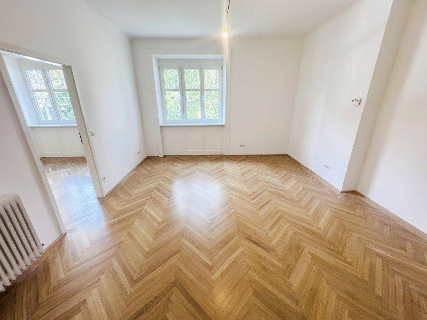 Geräumiges Zimmer mit elegantem Fischgrätparkett, einem Fenster und einem Heizkörper an der Wand.