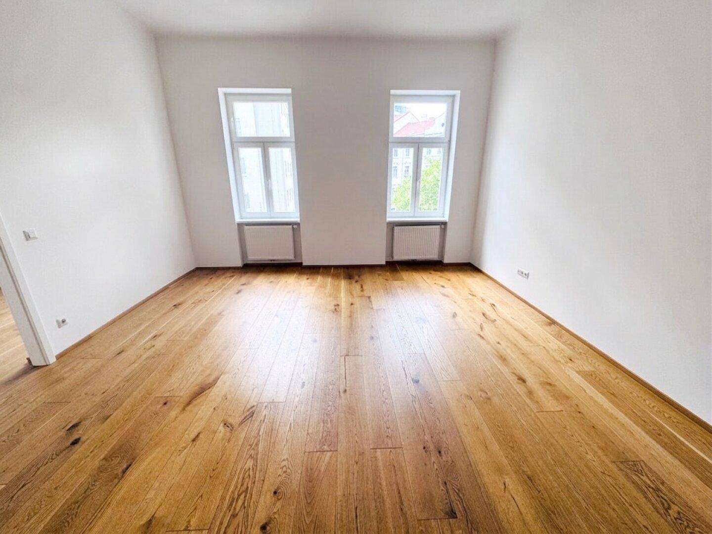 Geräumiger, leerer Raum mit hellem Holzboden und zwei hohen Fenstern, ideal für individuelle Gestaltung.