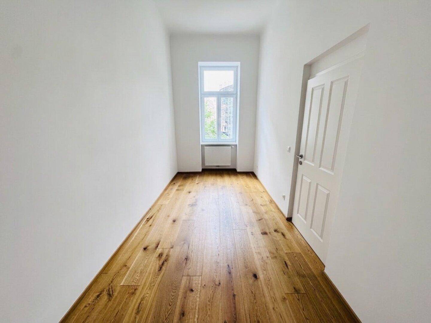 Langer, heller Raum mit Holzboden und einem Fenster, ideal als Schlafzimmer oder Arbeitszimmer.