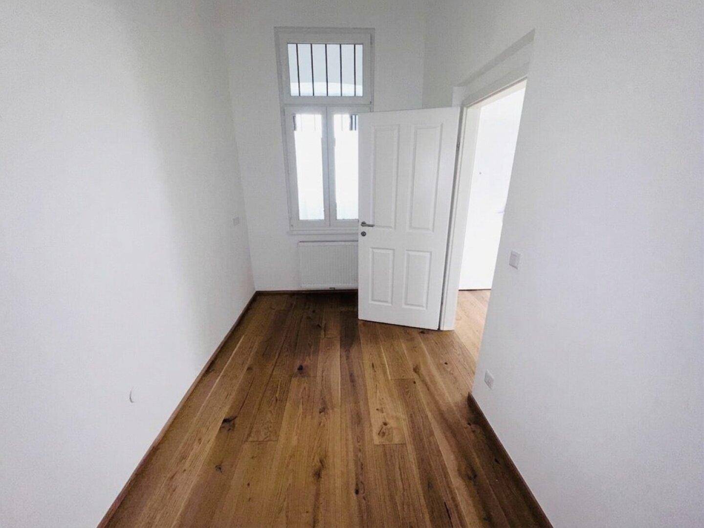 Schmaler, heller Raum mit Holzboden und einem Fenster, geeignet als Kinderzimmer oder Büro.