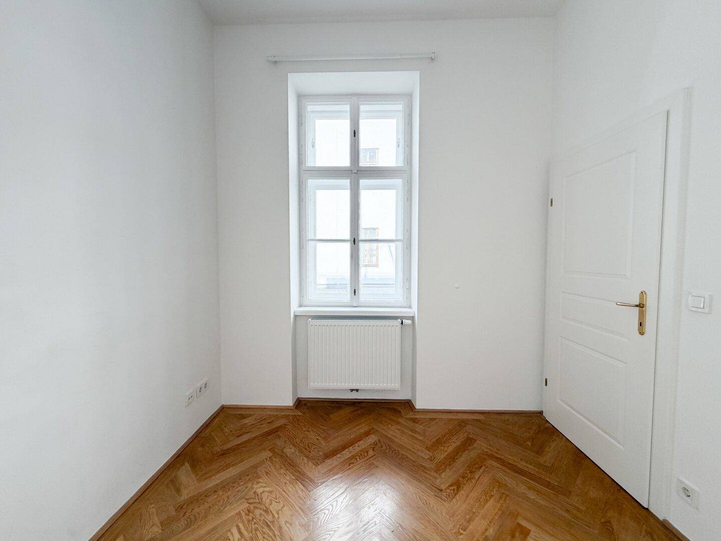 Ruhiges Zimmer mit Fischgrätparkett, einem Fenster und weißer Tür, ideal als Schlafzimmer.