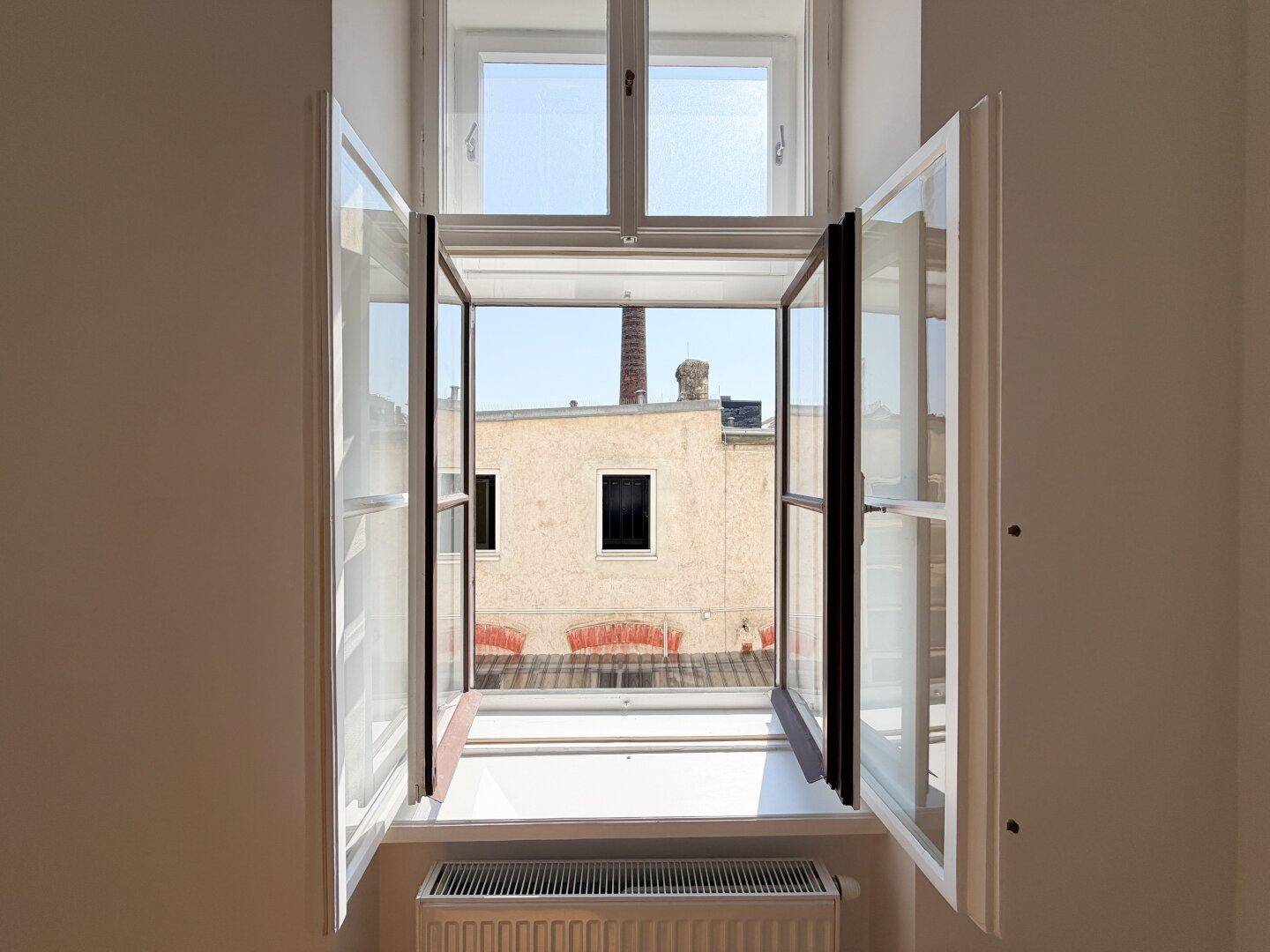 Blick aus dem geöffneten Fenster auf die umliegenden Gebäude und einen Schornstein.