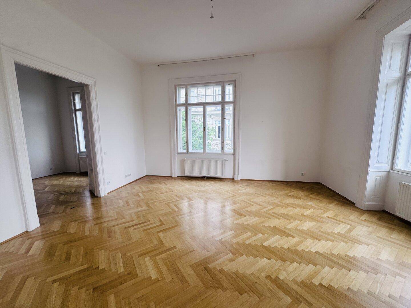 Geräumiges Zimmer mit hellem Fischgrätparkett, weißen Wänden und einem großen Fenster.