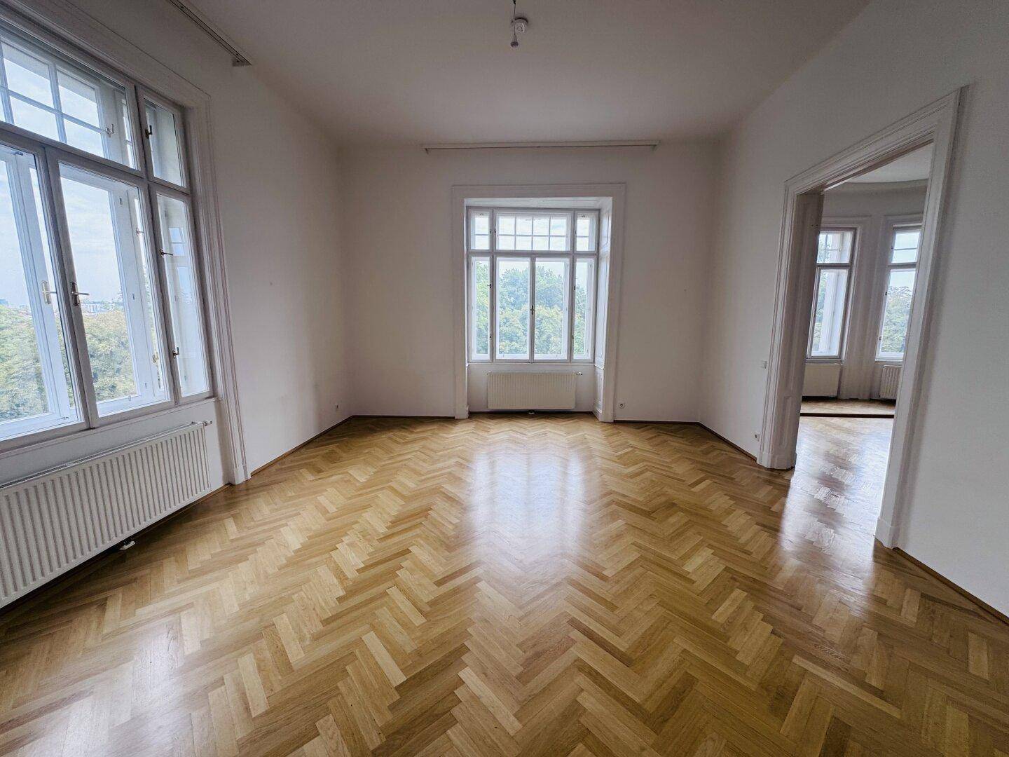 Helles Zimmer mit klassischem Fischgrätparkett, hohen Fenstern und Zugang zu weiteren Räumen.