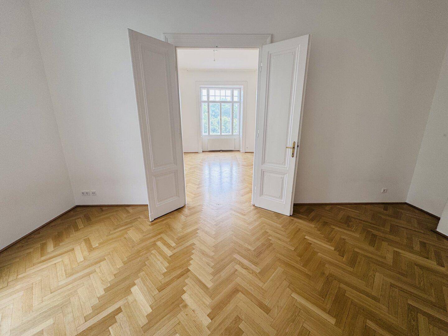 Blick durch eine offene Doppeltür in ein weiteres Zimmer mit klassischem Fischgrätparkett.