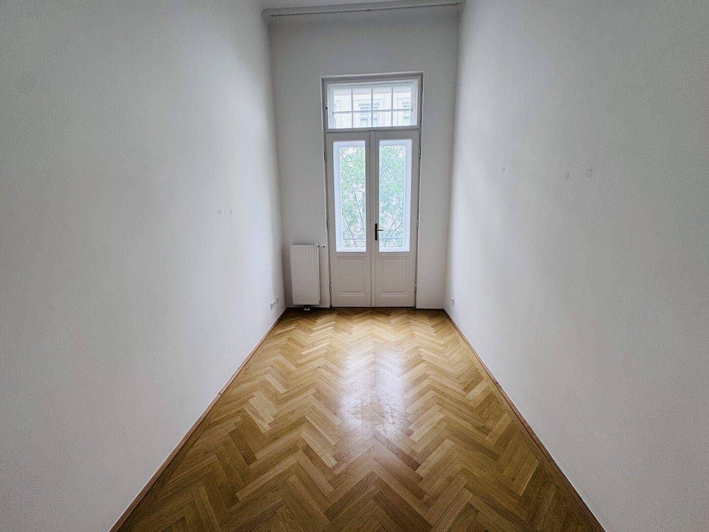 Schmaler Raum mit Fischgrätparkett und einer französischen Balkontür, die viel Licht hereinlässt.