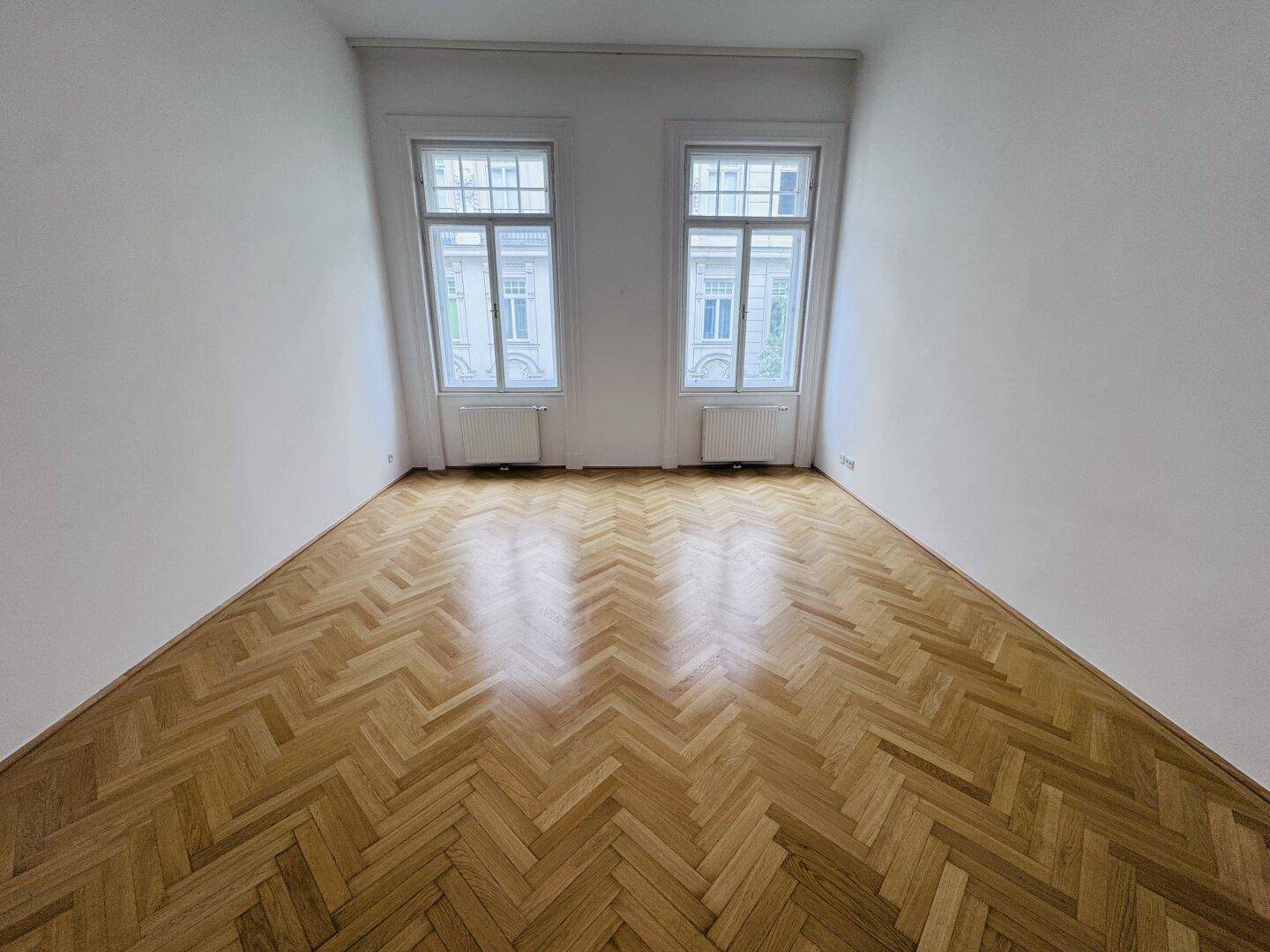 Ruhiges Zimmer mit klassischem Fischgrätparkett und zwei hohen Fenstern.