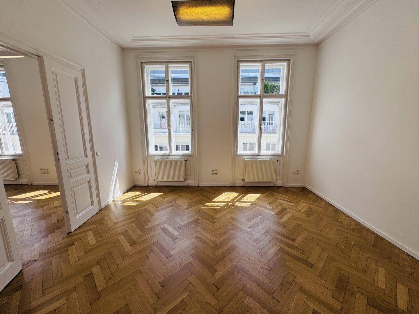 Geräumiges Zimmer mit Fischgrätparkett, hohen Decken und einem quadratischen Deckenlicht, das den Raum erhellt.