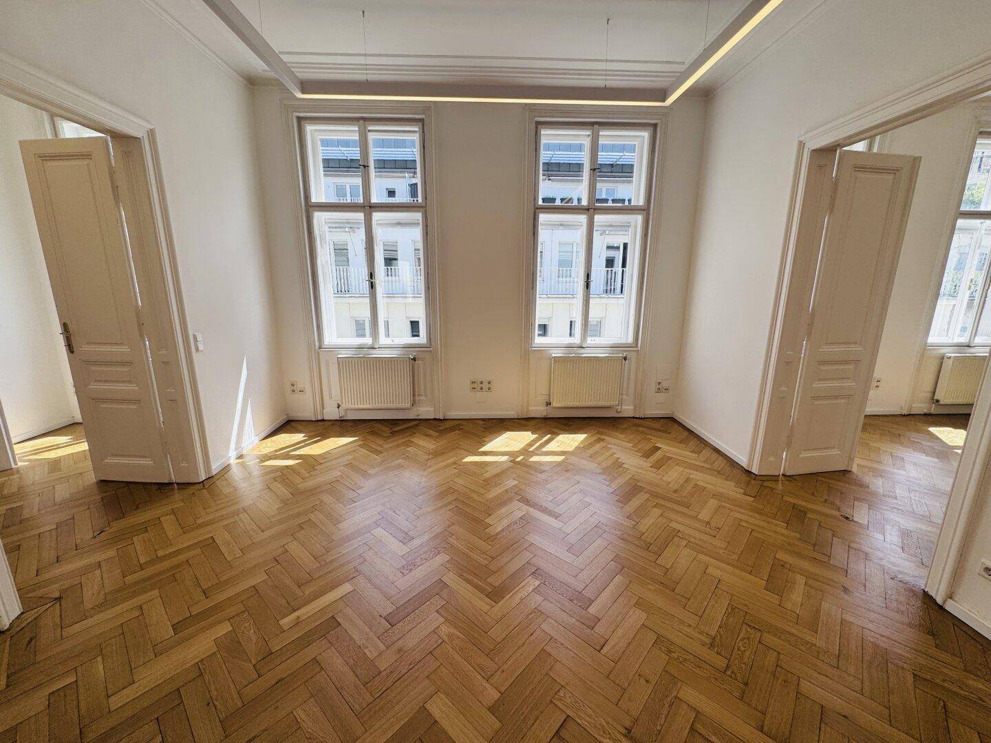 Geräumiges Zimmer mit Fischgrätparkett, hohen Decken und großen Fenstern, die viel Tageslicht hereinlassen.