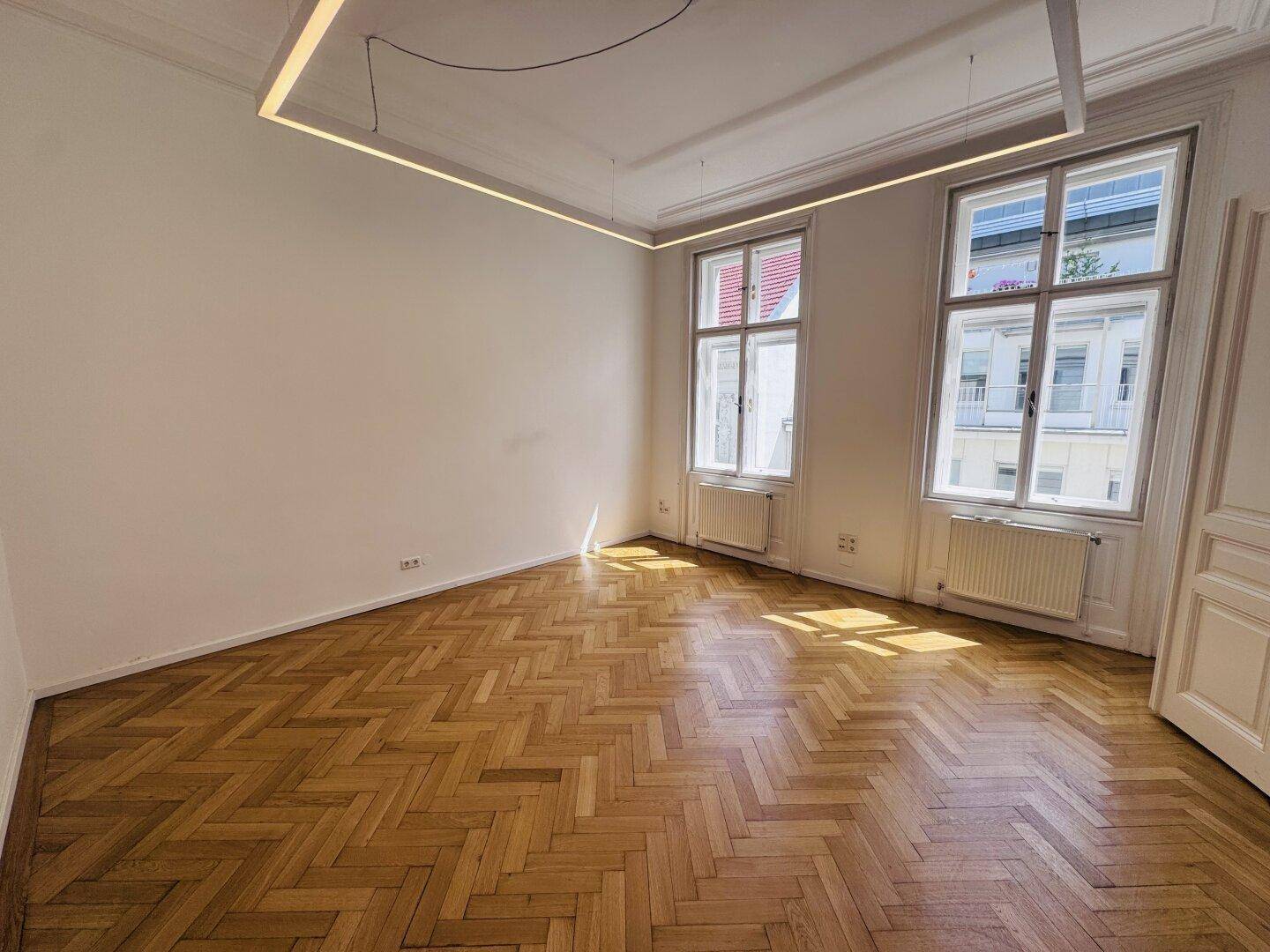 Helles Zimmer mit Fischgrätparkett und hohen Decken, durchflutet von natürlichem Licht durch große Fenster.