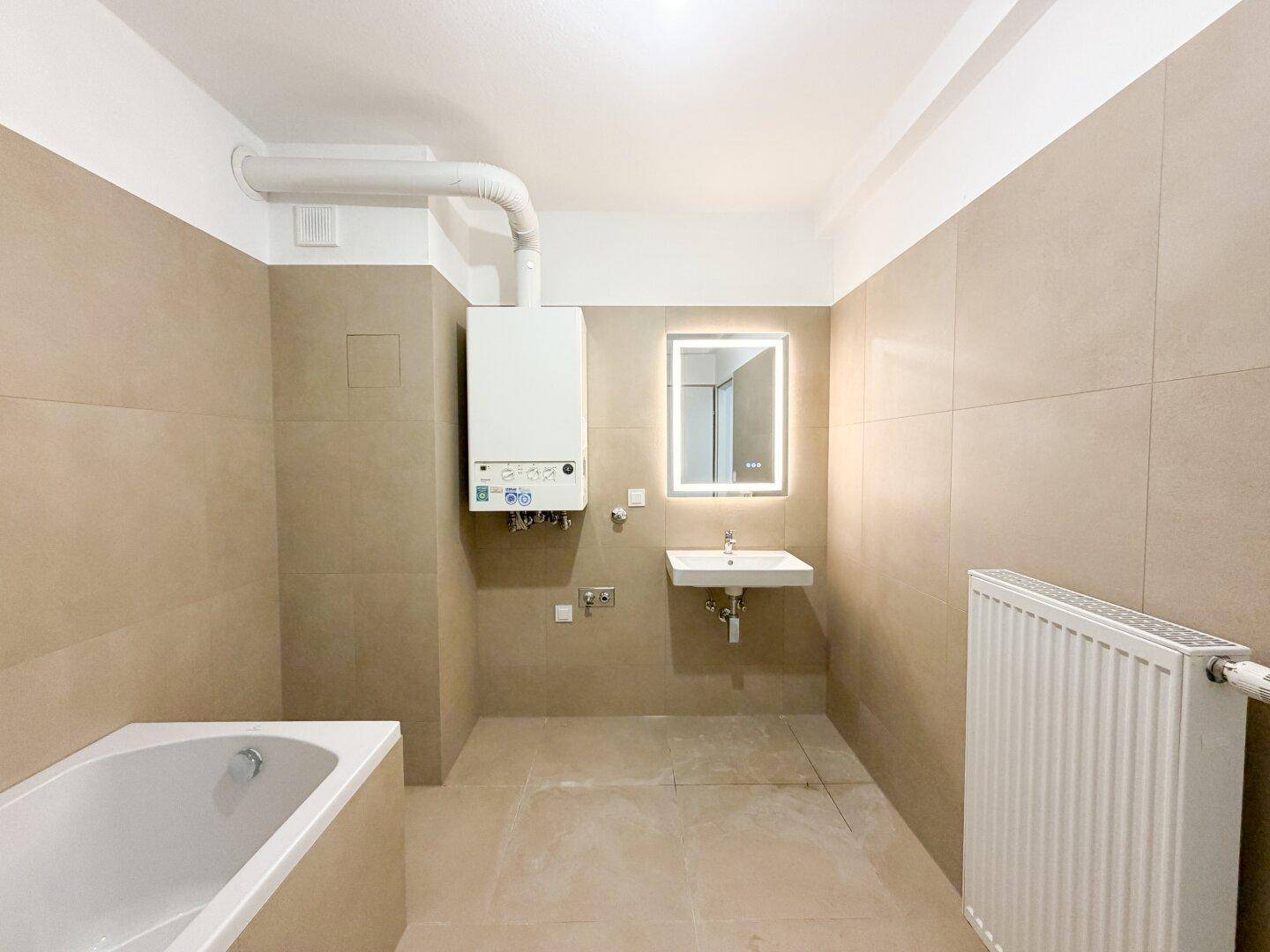 Helles Badezimmer mit Badewanne, Waschbecken, beleuchtetem Spiegel und Heizungsanlage.