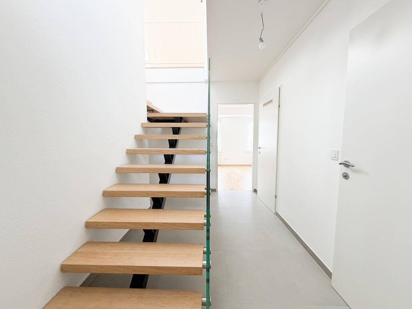 Heller Flur mit eleganter Holztreppe und Glasgeländer, führt zu weiteren Räumen.