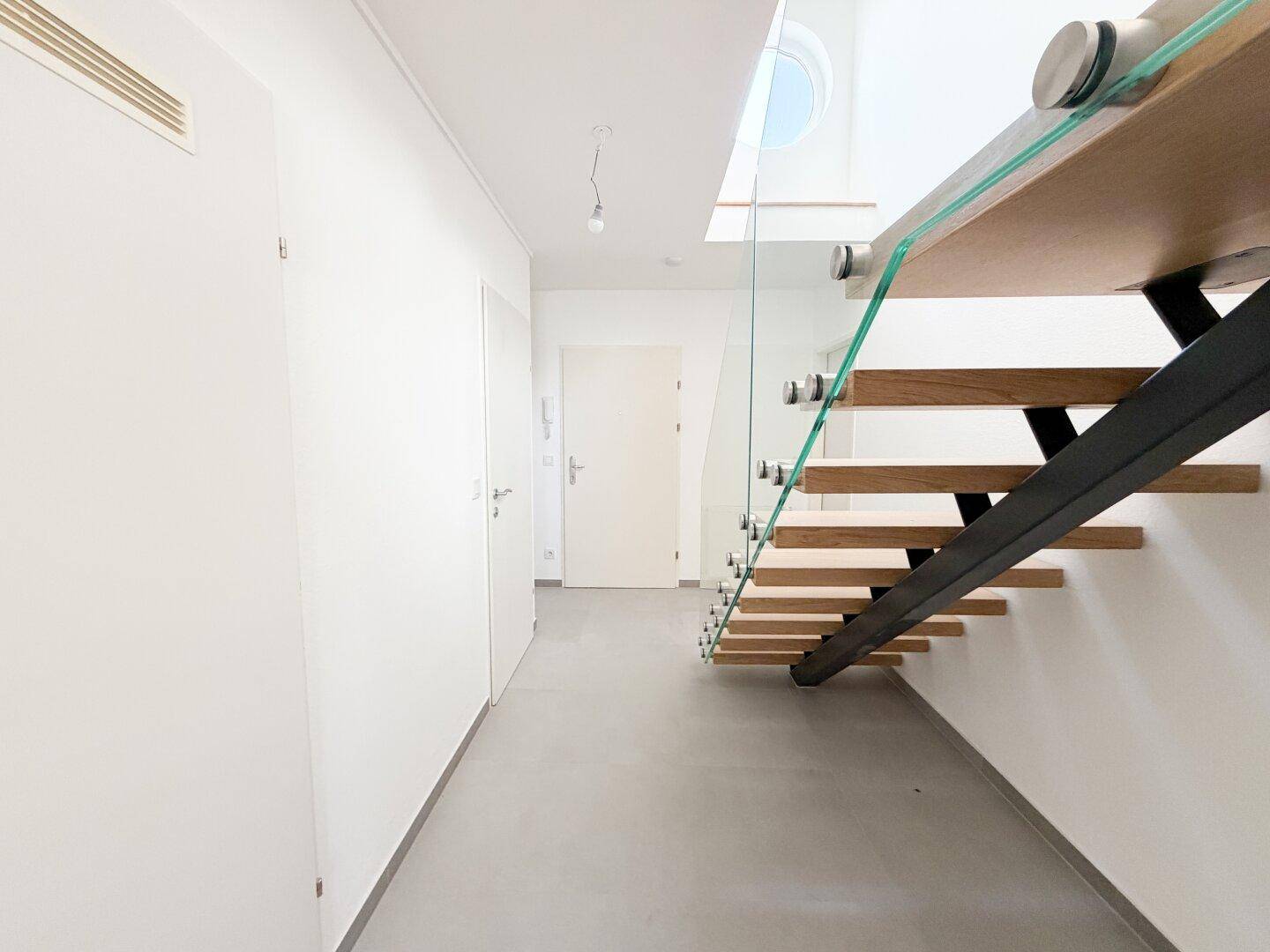 Moderner Flur mit schwebender Holztreppe, Glasgeländer und runden Fenstern.