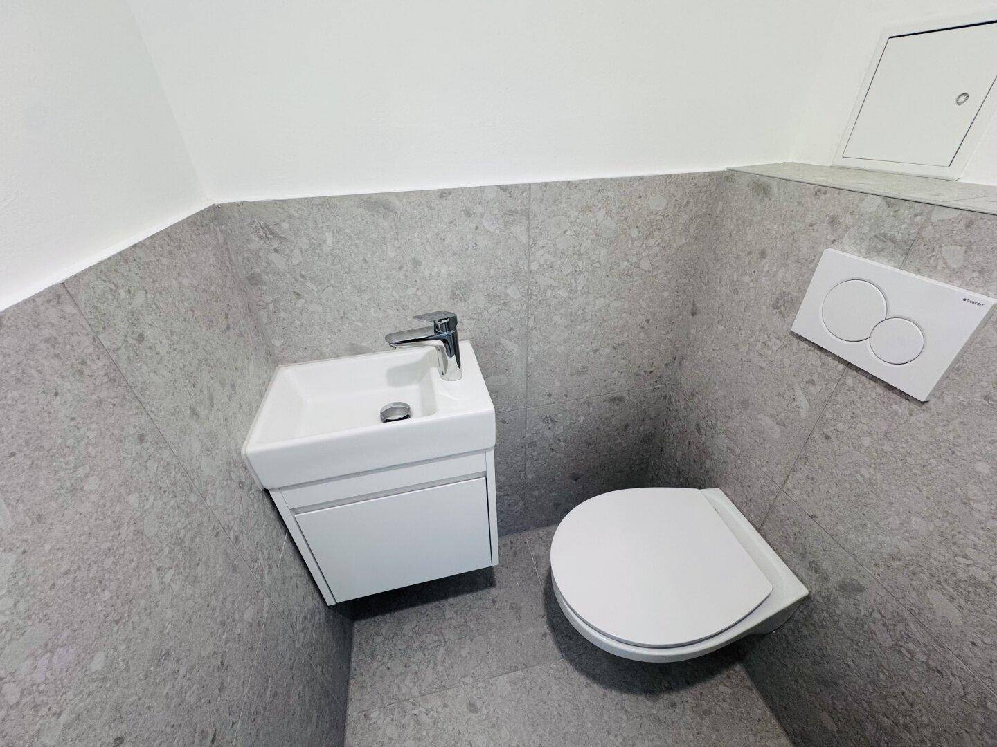 Modernes Gäste-WC mit wandhängender Toilette, kleinem Waschbecken und grauen Fliesen.