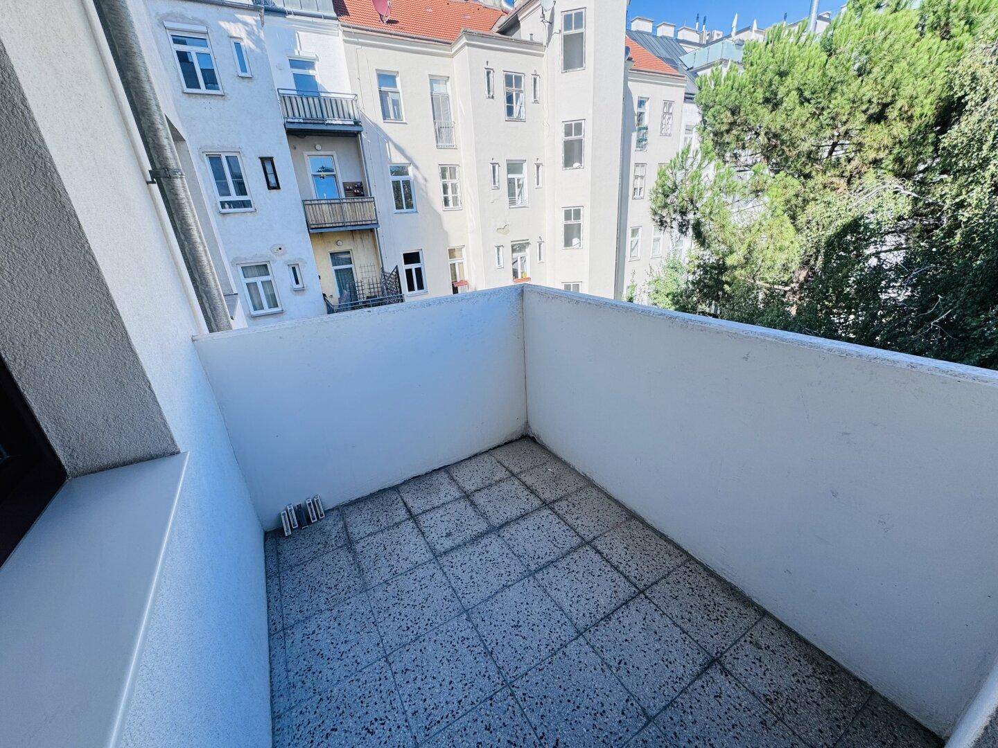 Leerer Balkon mit grauen Fliesen und weißen Wänden, bietet einen Blick auf die Nachbarschaft und Bäume.
