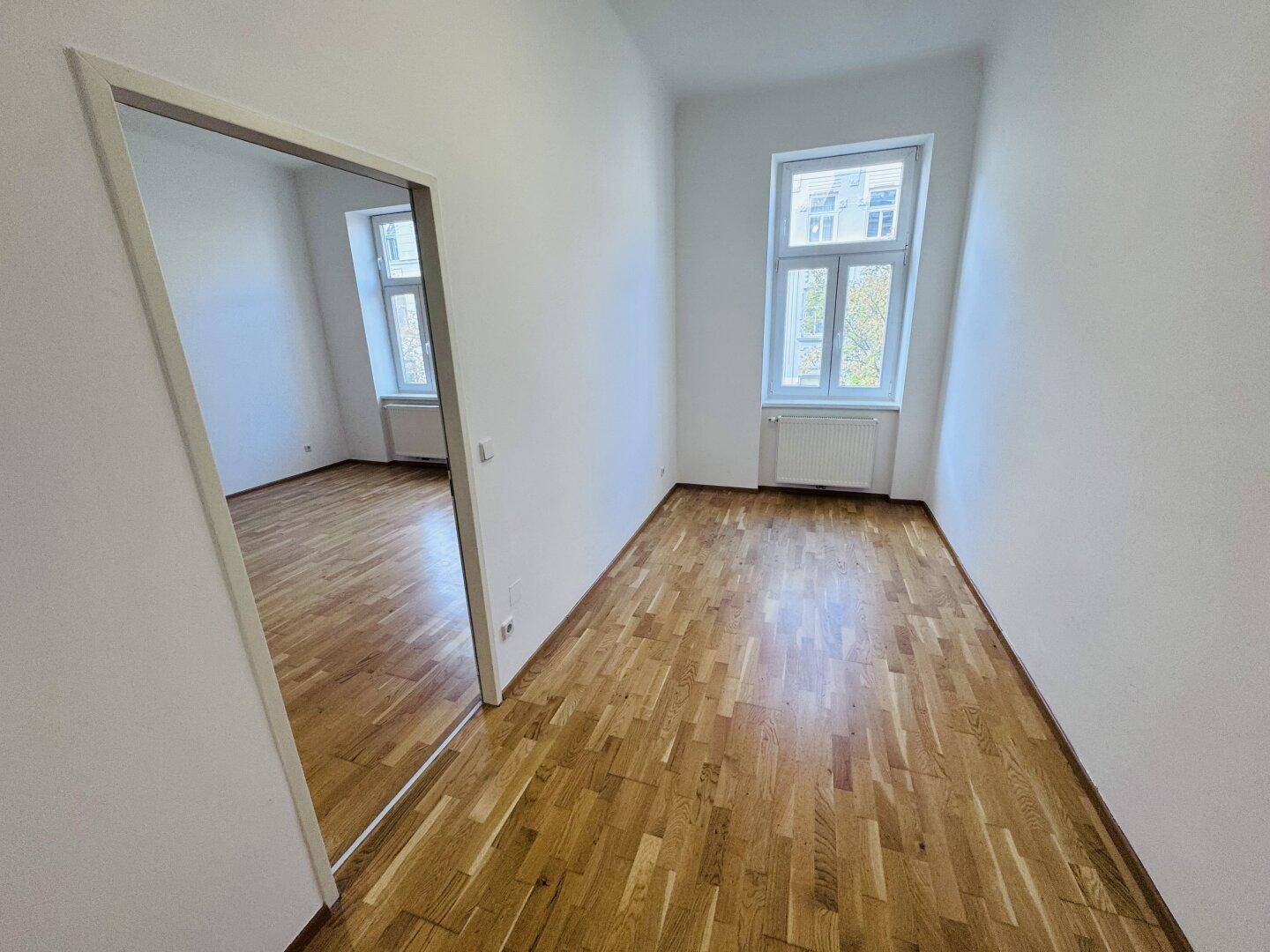 Geräumiges Zimmer mit Holzboden und hohen Decken, durch die Tür ist ein weiterer Raum sichtbar.