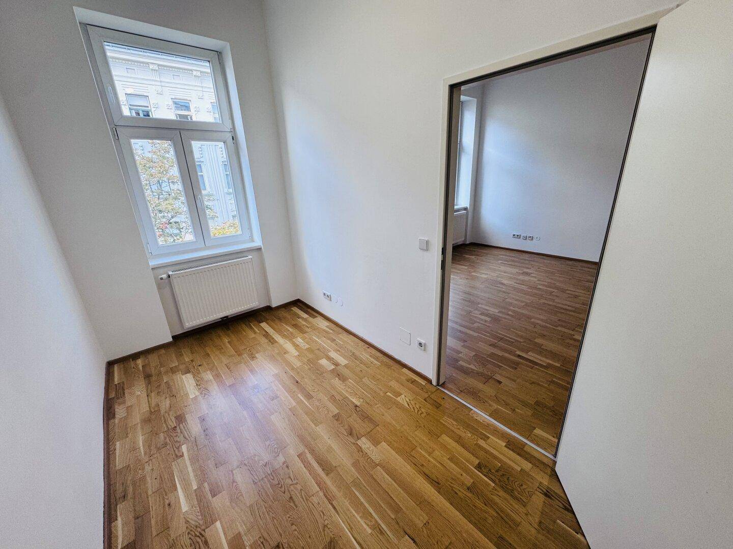 Heller Raum mit Parkettboden und einem großen Fenster, das viel Tageslicht hereinlässt.