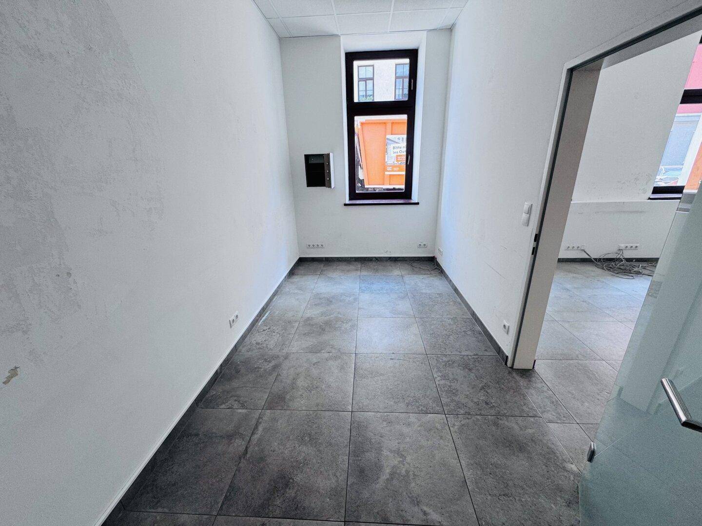 Kleiner Gewerberaum mit grauen Fliesen, weißen Wänden und einem Fenster zur Straße.