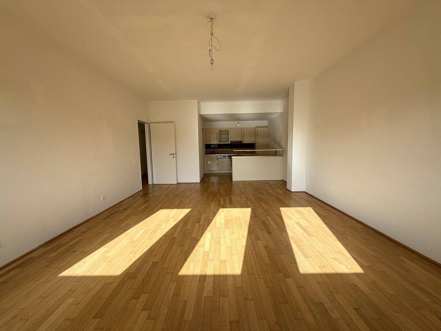 Heller, leerer Raum mit Holzboden und Übergang zu einem Küchenbereich im Hintergrund.